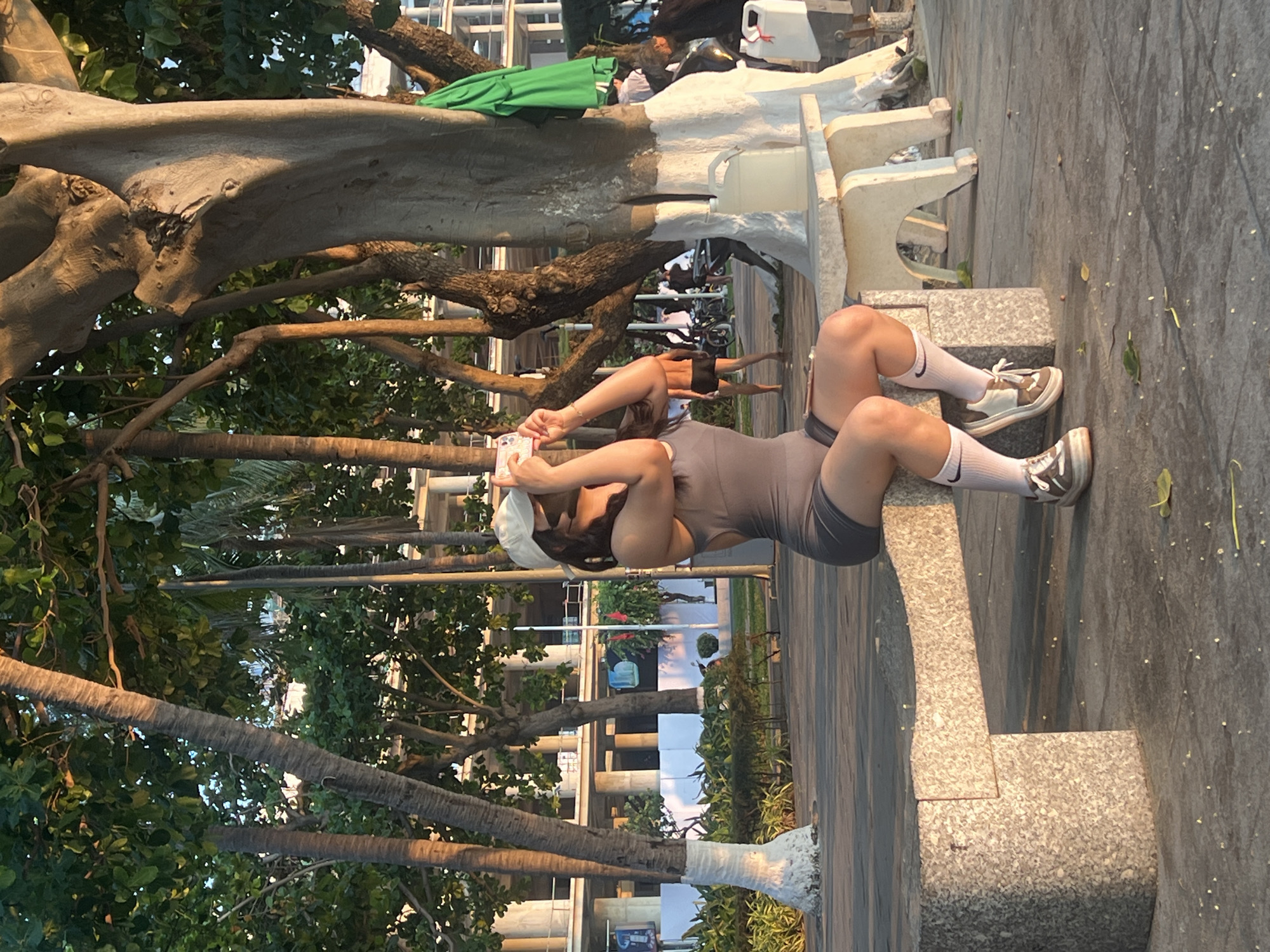 The image, taken in Nha Trang, Vietnam, captures an outdoor scene featuring a woman relaxing on a stone bench under the shade of trees. The woman is positioned on her back, wearing a form-fitting grey athletic bodysuit, a white baseball cap, white socks with the black Nike swoosh logo, and chunky white and brown athletic shoes. She holds a light-colored smartphone with both hands, positioned over her head.
The stone bench where she rests has a light, speckled grey surface. The ground immediately around the bench is a paved, light grey path, with a few scattered small leaves. Numerous trees frame the scene, including large trees with thick, gnarled trunks and branches, some of which have been painted white at their bases. Palm trees are also visible in the mid-ground and background, indicating a tropical or subtropical environment. Green foliage from various trees creates a natural canopy and backdrop.
In the background, other people are visible, including one person near bicycles and more distant figures, suggesting a public park or promenade. A green fabric object is draped over a tree branch, and a white plastic container sits on a raised structure. Modern buildings are discernible through the trees in the far background.
The lighting indicates either late afternoon or early morning, with warm, soft sunlight illuminating parts of the scene and casting long shadows, suggesting clear weather. The overall atmosphere is relaxed and peaceful, indicative of leisure time in an urban green space. The "Nike" logo on the woman's socks is the only visible text.