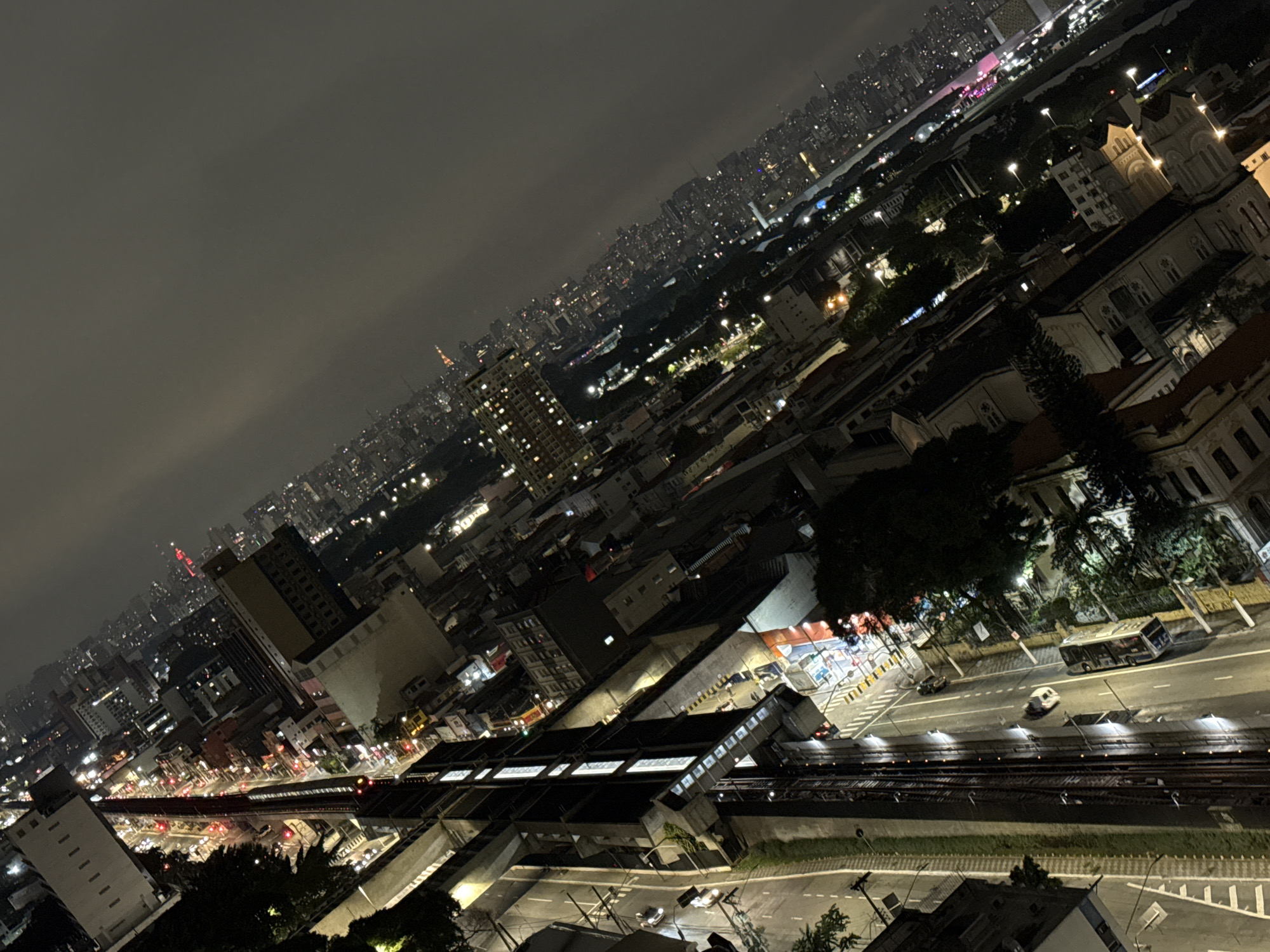 The image depicts an elevated nighttime cityscape in São Paulo, Brazil, under an overcast sky. City lights illuminate densely packed buildings extending into the distance. In the foreground, a well-lit train station with tracks runs parallel to a street with buses and cars. Colonial architecture stands near the train station. People cannot be distinguished.