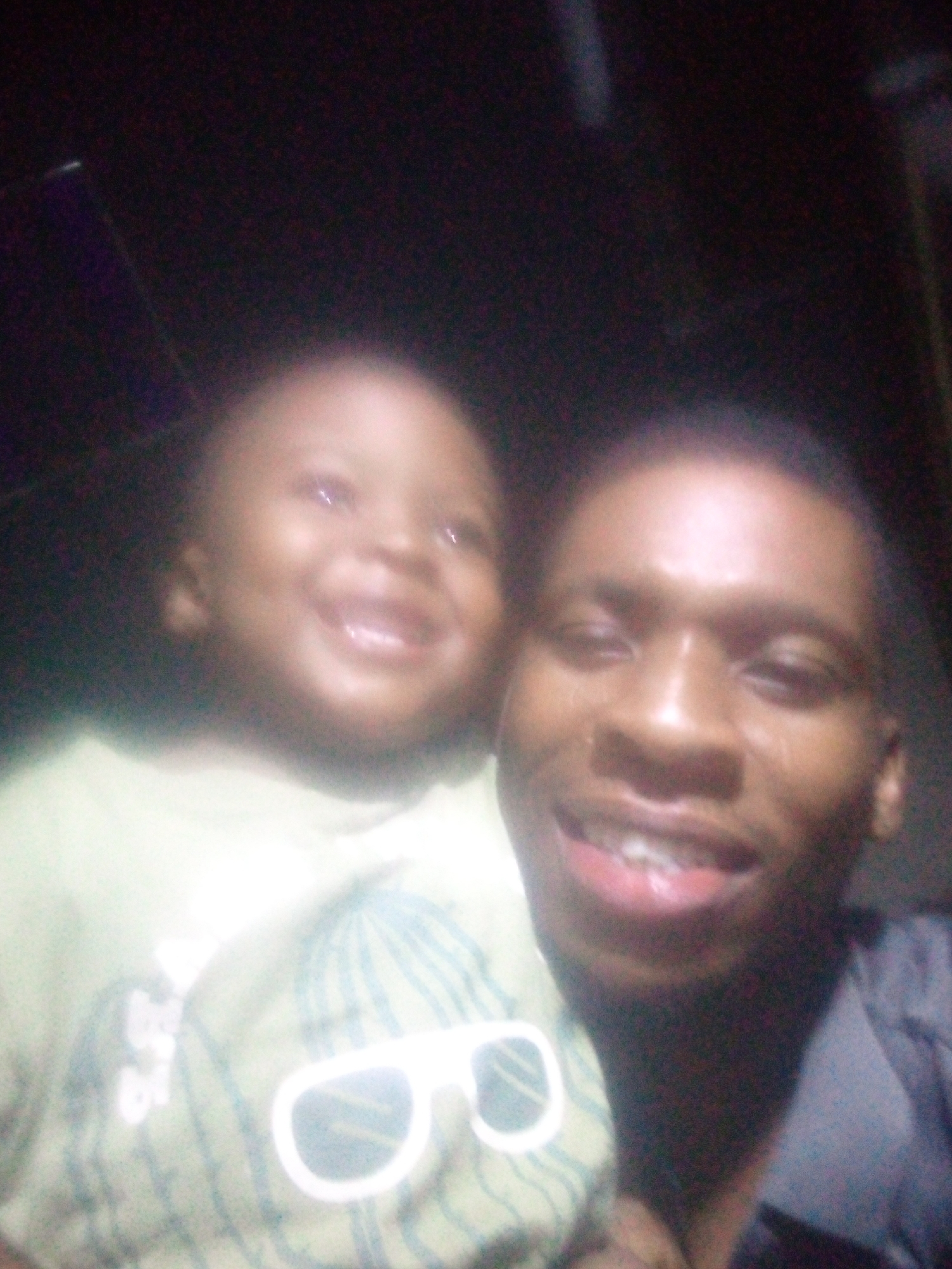 A close-up, blurry, and grainy image shows two individuals, an adult male and a young child, posing together, likely in a selfie. The lighting is very dim, with a bright flash or light source illuminating their faces, causing some overexposure and lens flare.
The adult male, on the right, has dark skin and short dark hair. He is smiling widely, revealing his teeth, and his eyes are open. He appears to be wearing a dark, collared shirt.
The child, on the left, is positioned slightly behind and to the left of the adult. The child also has dark skin and is smiling broadly, showing a few teeth. The child wears a light-colored t-shirt with a graphic design. The design features a large, stylized pair of white sunglasses with thick frames, positioned over what might be a patterned hat (possibly striped or checkered). To the left of the sunglasses design, some blurry white text is visible, but too indistinct to read clearly.
The background is almost entirely black and featureless, indicating very low ambient light. Both subjects appear happy and joyful, with prominent smiles. The overall image quality is low, characterized by noise, blur, and strong contrast due to the localized light source. No specific location or time of day beyond "low light" can be determined.