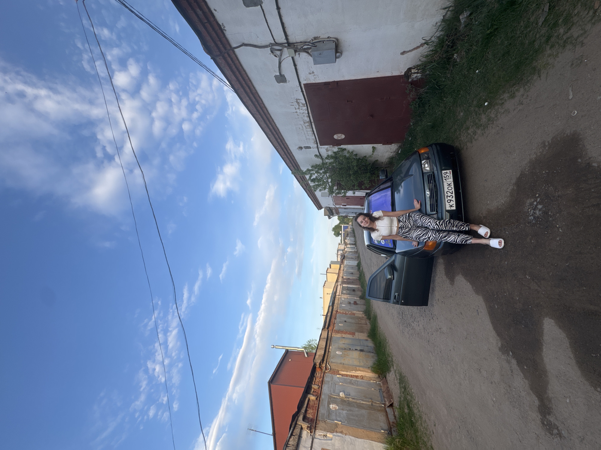 A woman lies on the windshield of a dark green Lada car, license plate number K932OK159, on a dirt road between rows of garages. The woman wears a white top, zebra-print leggings, and white slides. The location appears to be an alley of garages in Chaikovsky, Russia. The sky is blue with scattered clouds. Overhead power lines are visible.
