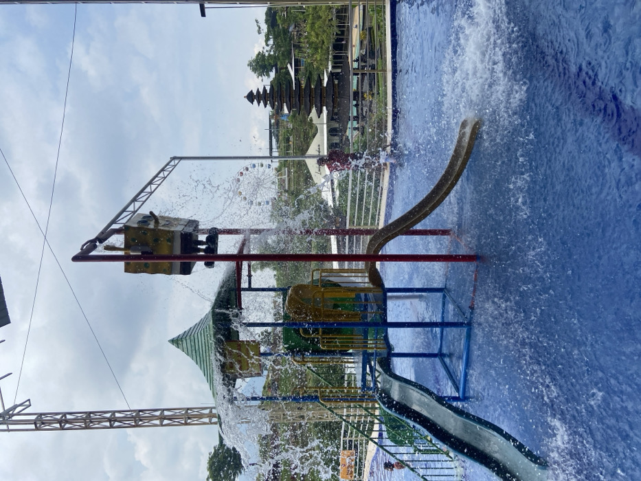 A vibrant water park scene features a colorful play structure with slides and a large bucket pouring water. In the background, a towering pagoda-style building and a Ferris wheel are visible. The water park has a blue tiled base, and trees are scattered around the park. Water droplets are captured mid-air, adding a dynamic feel to the image. The sky is partly cloudy.