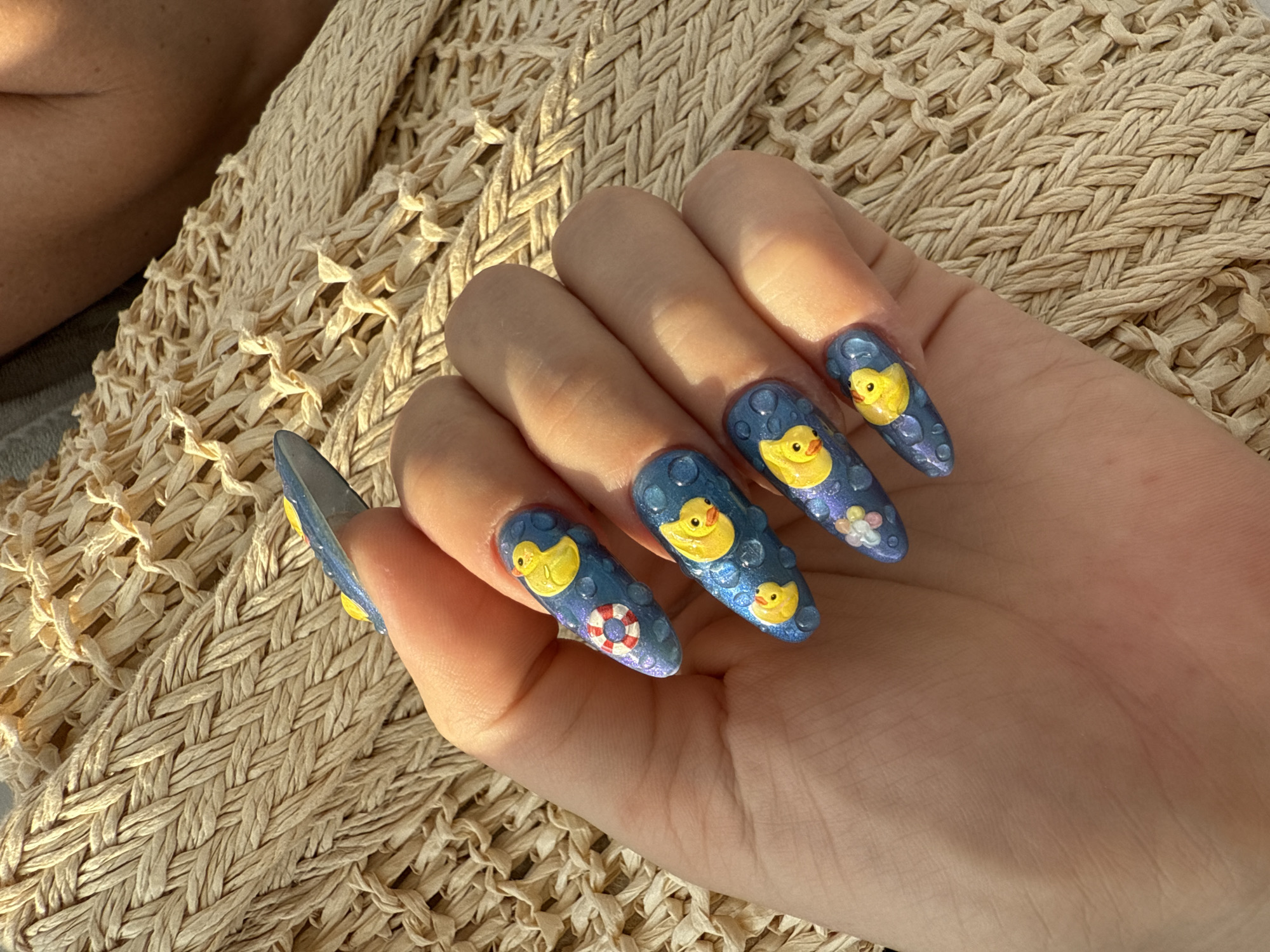 A close-up overhead shot shows a woman's hand with elaborately decorated fingernails against a textured, woven, light-beige background. The nails are long and almond-shaped, painted a shimmery blue with raised 3D designs. Each nail features at least one yellow rubber duck. Some nails also have a red and white life preserver ring, or small floral designs. The blue nail polish has small raised, clear bumps that resemble water droplets. The background appears to be a woven mat or fabric, possibly a beach towel or a sun lounger, with a person's skin visible in the upper right corner.