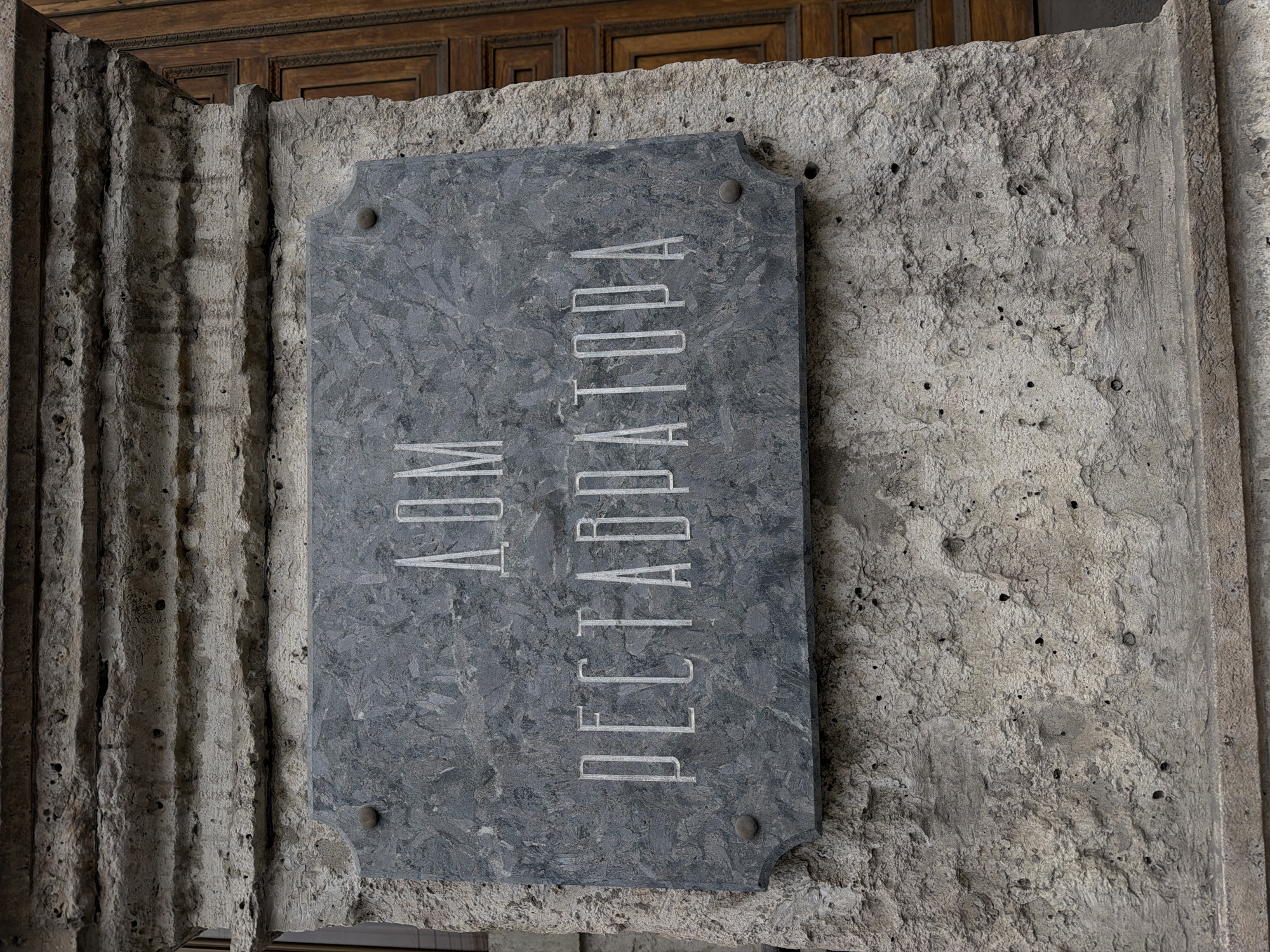 A stone plaque bearing the Cyrillic inscription "ДОМ ПЕГРАДАТОРА" is affixed to a textured stone wall. The plaque, a dark gray marble-like material, is rectangular with rounded corners secured by four metal fasteners. The inscription is oriented vertically and appears to be engraved or etched. The stone wall has a rough, porous texture. A fluted stone column or structure appears to the left of the plaque, and part of a wooden door is visible at the top of the frame. The setting suggests a historical or architectural context within the city of Saint Petersburg, Russia.