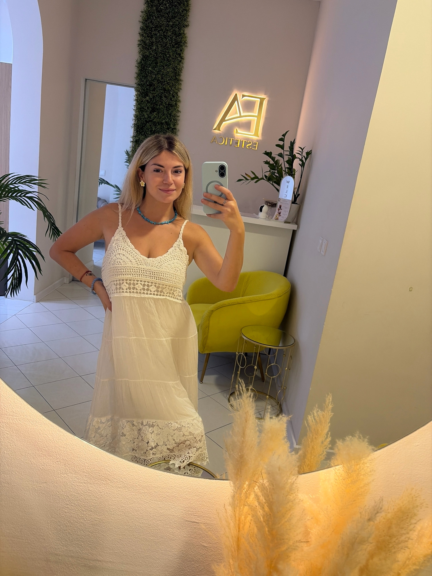 A young blonde woman with blue eyes is taking a mirror selfie in what appears to be a modern beauty salon in Budva, Montenegro. She is wearing a white crocheted and tiered maxi dress, a turquoise beaded necklace, and a flower-shaped earring. She is also wearing bracelets on her left wrist. In the reflection of the mirror, a yellow armchair, a small golden table with a glass top, and a decorative plant are visible. Behind the woman, a neon sign that reads "EA ESTETICA" is illuminated on the wall. The floor is tiled with white squares. The overall environment suggests a chic and clean establishment.