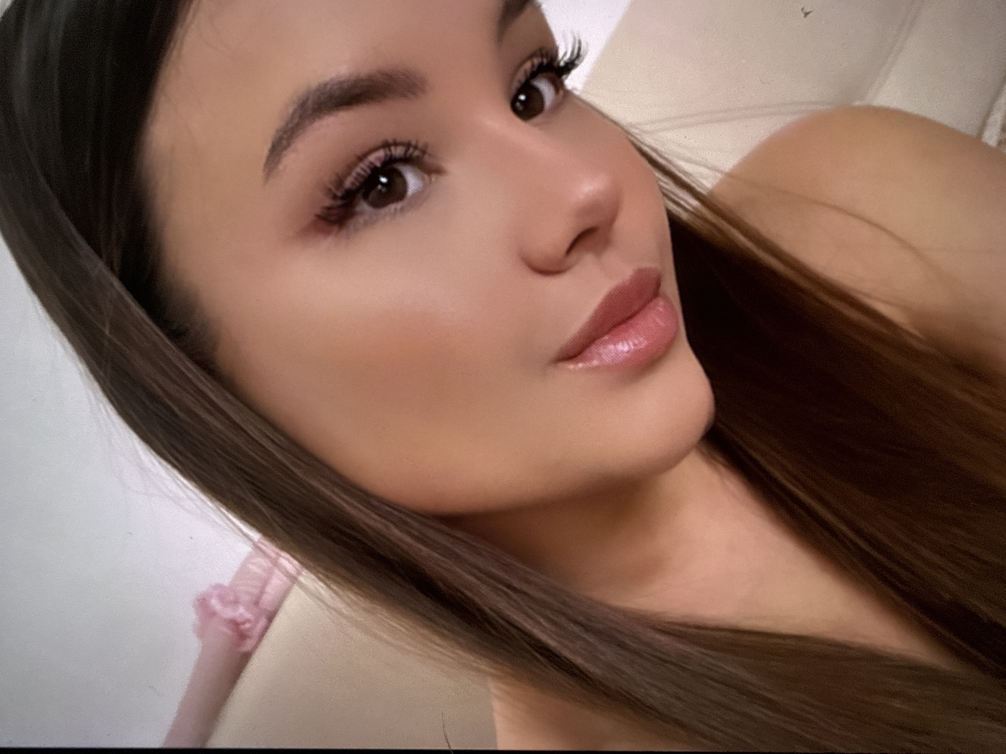 A close-up, angled headshot of a young woman with light brown eyes and dark brown hair. She has well-defined eyebrows and is wearing makeup, including eyelashes and lip gloss. A portion of her right shoulder is visible, along with a light-colored background with a hint of pink fabric on the left side of the image. There are no indicators of the location being Sofia, Bulgaria, or any city. There is no action, interaction, or indication of environment.