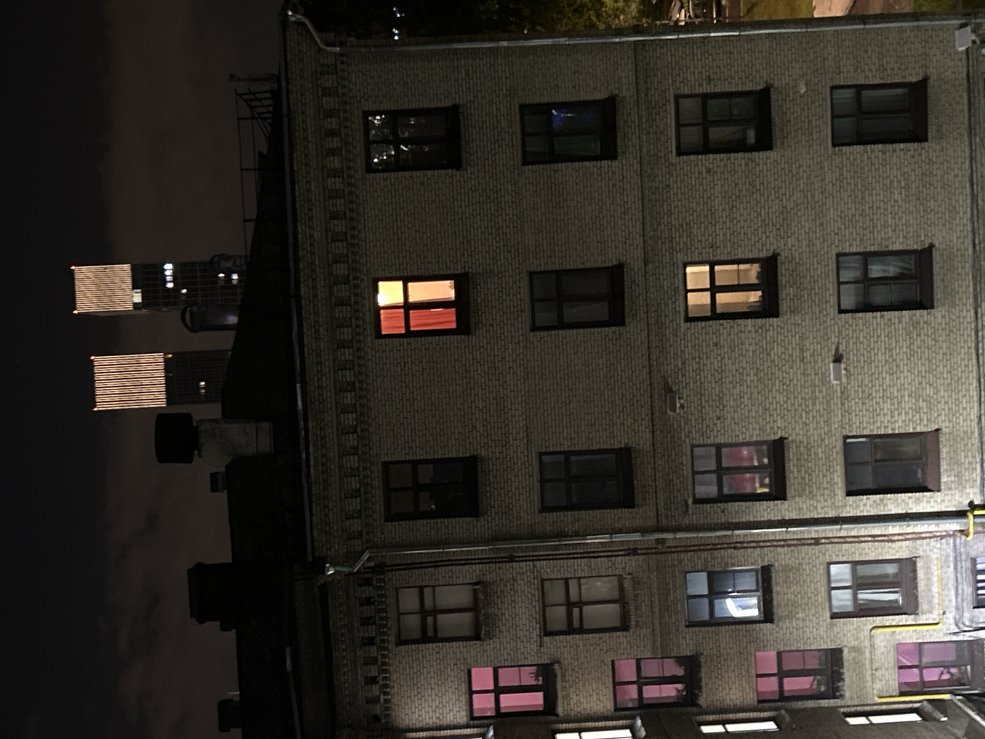 At night, the exterior of a building is illuminated with various lights from the windows. The brick facade of the building features an array of windows. Some windows are lit with a warm, orange glow, others display a pink hue, and some appear dark. In the background, the skyline is visible, with a couple of taller structures lit up. The overall scene is urban.