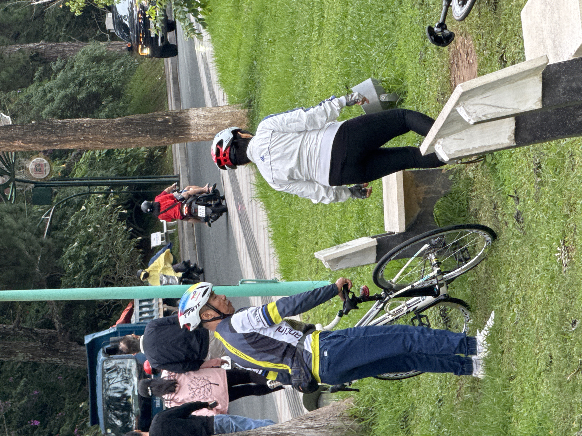 The image depicts a street scene in Da Lat, Vietnam, with a focus on individuals engaged in various activities. In the foreground, a man wearing a cycling jersey and helmet stands beside a bicycle. To his right, another person in a white jacket and black leggings, also wearing a helmet, walks away from the camera. The background reveals a road with passing vehicles, including a blue truck and a black SUV. Several people are seen on motorcycles, one of whom is wearing a yellow poncho. The environment consists of lush green grass, trees, and a light pole. The overall atmosphere suggests a casual outdoor setting, possibly a park or a roadside in Da Lat.