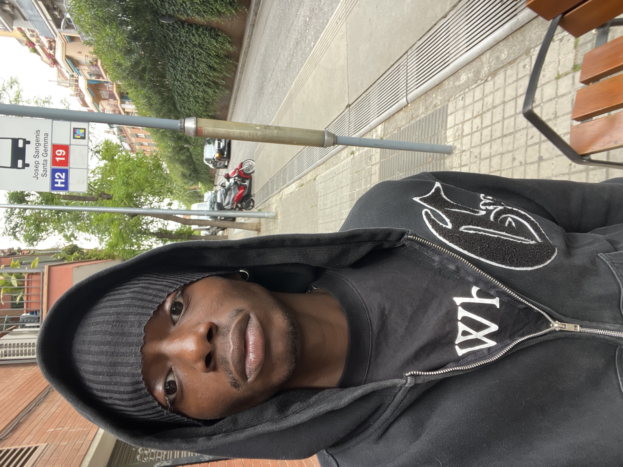 A close-up, canted shot shows a Black male wearing a black hooded sweatshirt with a white design and lettering. He also wears a black and grey striped beanie. He is situated at an outdoor bus stop, identifiable by the "Josep Sangenis Santa Gemma" sign featuring the bus route numbers H2 and 19. The setting is plausibly in Barcelona, Spain, given the language on the bus stop sign. A metal pole and part of a wooden bench are also visible. The background includes an urban street scene with buildings and vegetation.