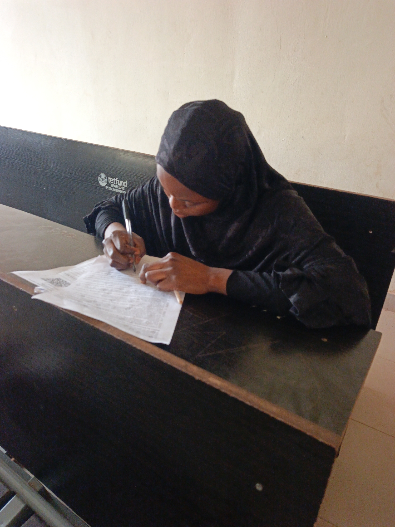 A woman wearing a black hijab is seated at a dark-colored desk, writing on a piece of paper with a black pen. The desk has a Tetfund logo on the back. The location is presumed to be in Akwanga, Nigeria, based on visual cues.