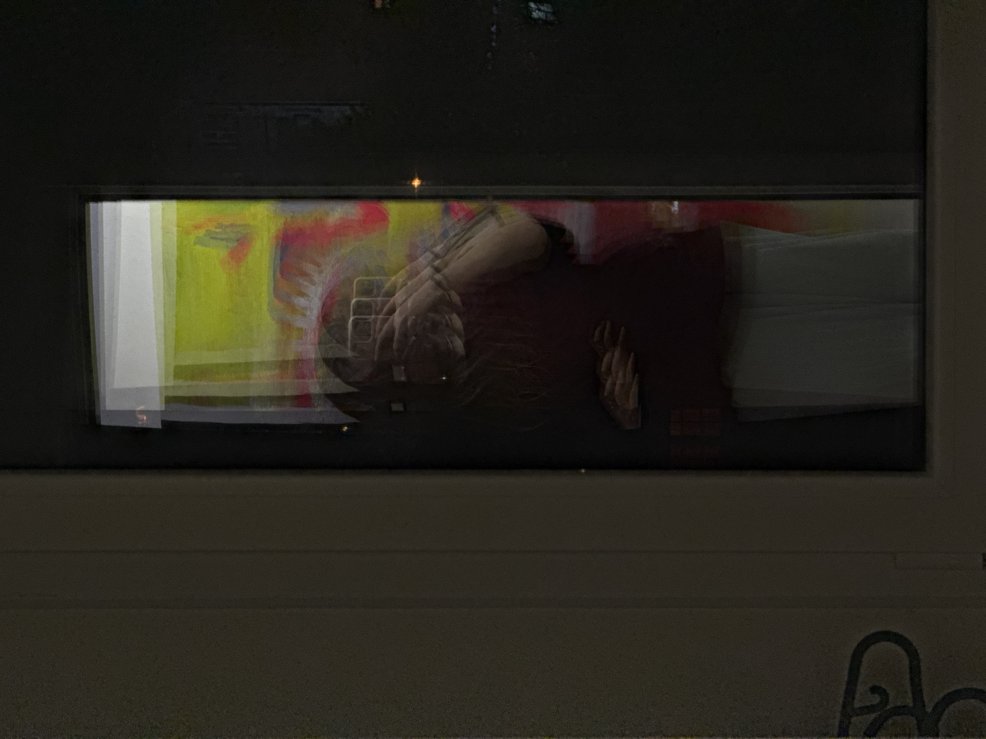 A blurred image shows a person in a dark room, reflected in what appears to be a glass surface or window. The person is wearing a dark-colored shirt. Behind the person is a yellow and red abstract background. The environment is dark, with a part of a surface visible in the lower part of the image. Based on the request for the location, the image is assumed to be taken in the city of Vozdovac Urban Municipality, Serbia.