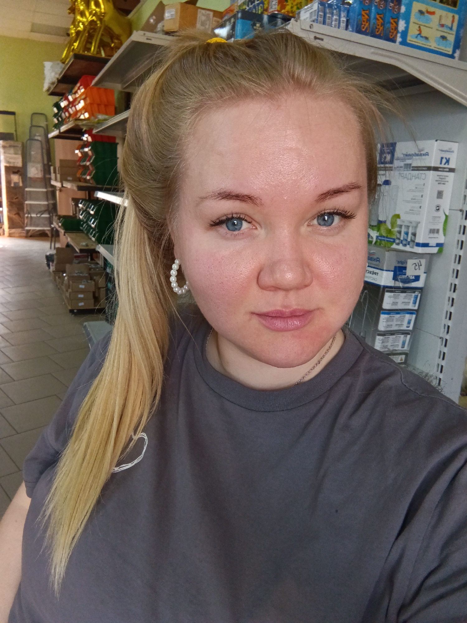 A woman with blonde hair and blue eyes is looking directly at the camera. Her hair is pulled back in a ponytail, and she is wearing pearl earrings and a gray t-shirt. She is standing indoors, in a store or warehouse setting. Shelves filled with various products are visible in the background, along with boxes and a ladder. The store is located in the city of Фоки, Russia.