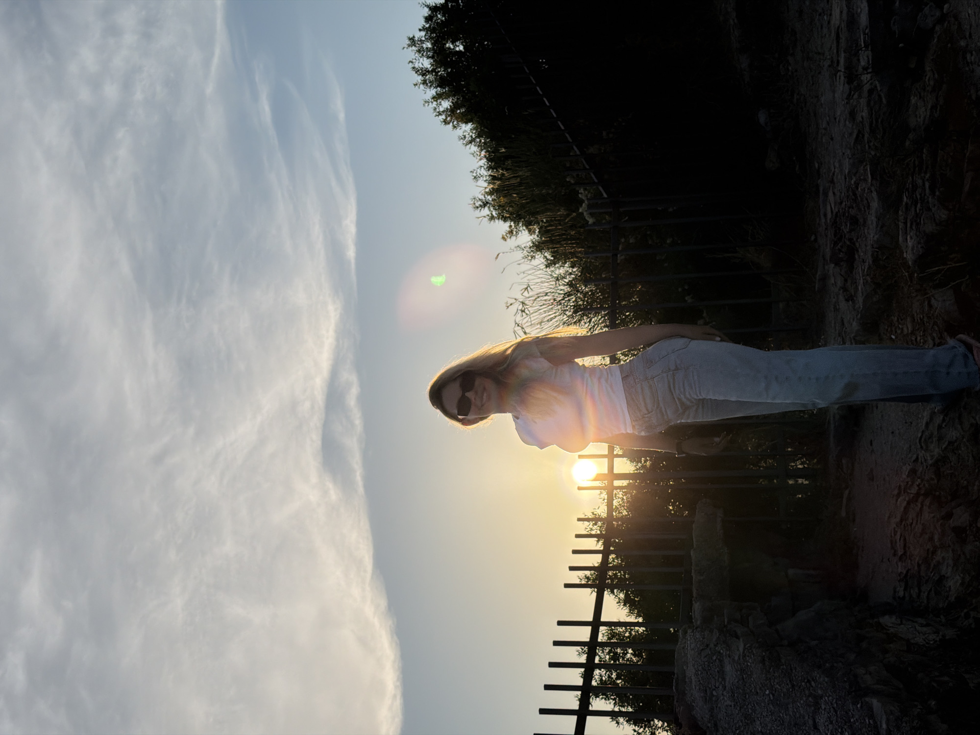 A young woman with blonde hair, wearing a white t-shirt, light blue jeans, and sunglasses, stands outdoors in what appears to be Budva, Montenegro. She is positioned in front of a black metal fence, with trees and foliage behind it. The sun is low in the sky, creating a warm, hazy light and a lens flare effect. The background consists of a gradient sky with wispy clouds, transitioning from a pale yellow near the horizon to a soft blue. The foreground shows rocky terrain.