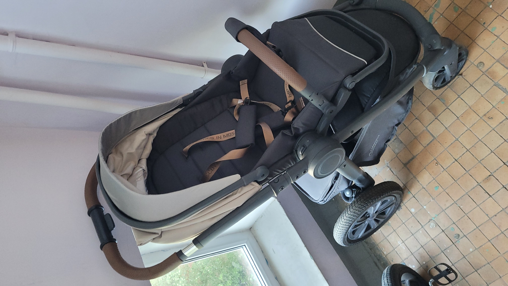 The image shows a stroller indoors, placed near a window and a wall. The stroller is dark grey with brown leather accents on the handle and straps. The floor is covered with square terracotta tiles, some of which have green stains. There is also a white pipe running horizontally along the wall near the top of the image. A blurred view through the window is visible, suggesting an outdoor scene.