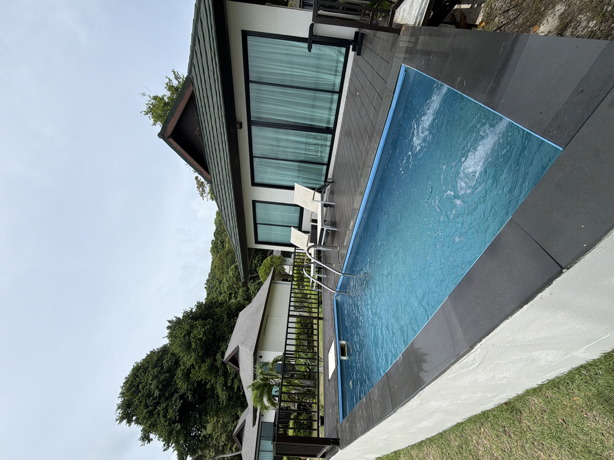 The image depicts a private swimming pool and a modern villa exterior, likely a resort, in Ao Nang, Thailand. The rectangular pool, filled with clear blue water, features bubbling jets and is bordered by a dark tiled coping. Two white lounge chairs with metal frames are positioned on a dark wooden deck adjacent to the pool. A white platform is visible within the pool's shallow end. The villa has a white facade, a dark tiled roof, and large sliding glass doors with sheer curtains. A dark metal railing encloses the deck area. In the background, lush green trees and other villa structures are visible, suggesting a tropical environment. The sky is overcast and pale. The scene is tranquil and suggests a luxurious vacation setting.