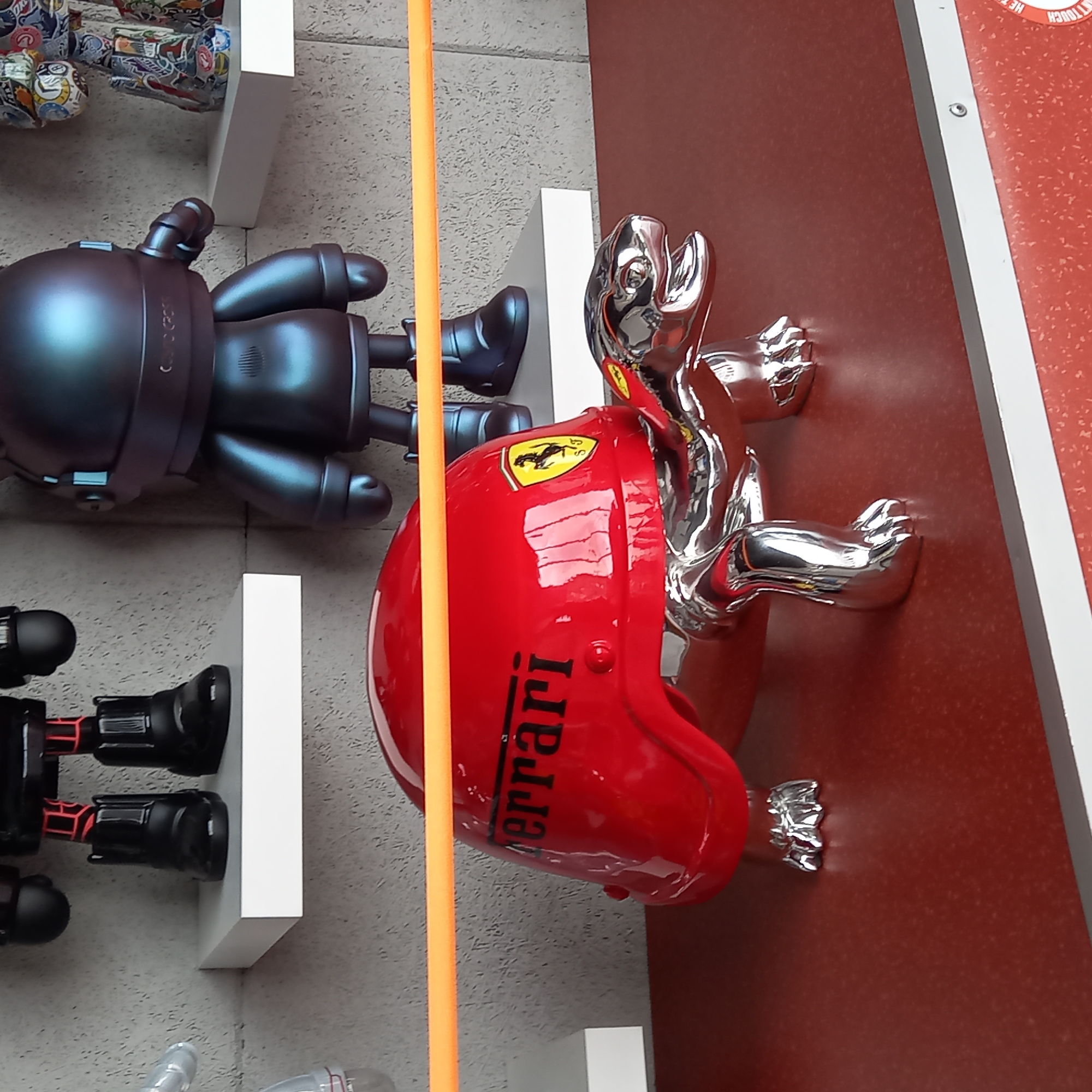 In a dimly lit indoor setting, a bright red Ferrari-themed turtle figurine is prominently displayed on a textured red surface. The turtle has a glossy red shell adorned with the Ferrari logo and lettering. Its body and limbs are made of polished chrome, reflecting the surrounding environment. Behind the figurine, on a grey, textured wall, are shelves holding other decorative items, including a black robot figurine and a more colorful, multi-patterned figure. An orange bar or strap extends horizontally across the image in front of the turtle. The overall scene appears to be a retail or display environment.