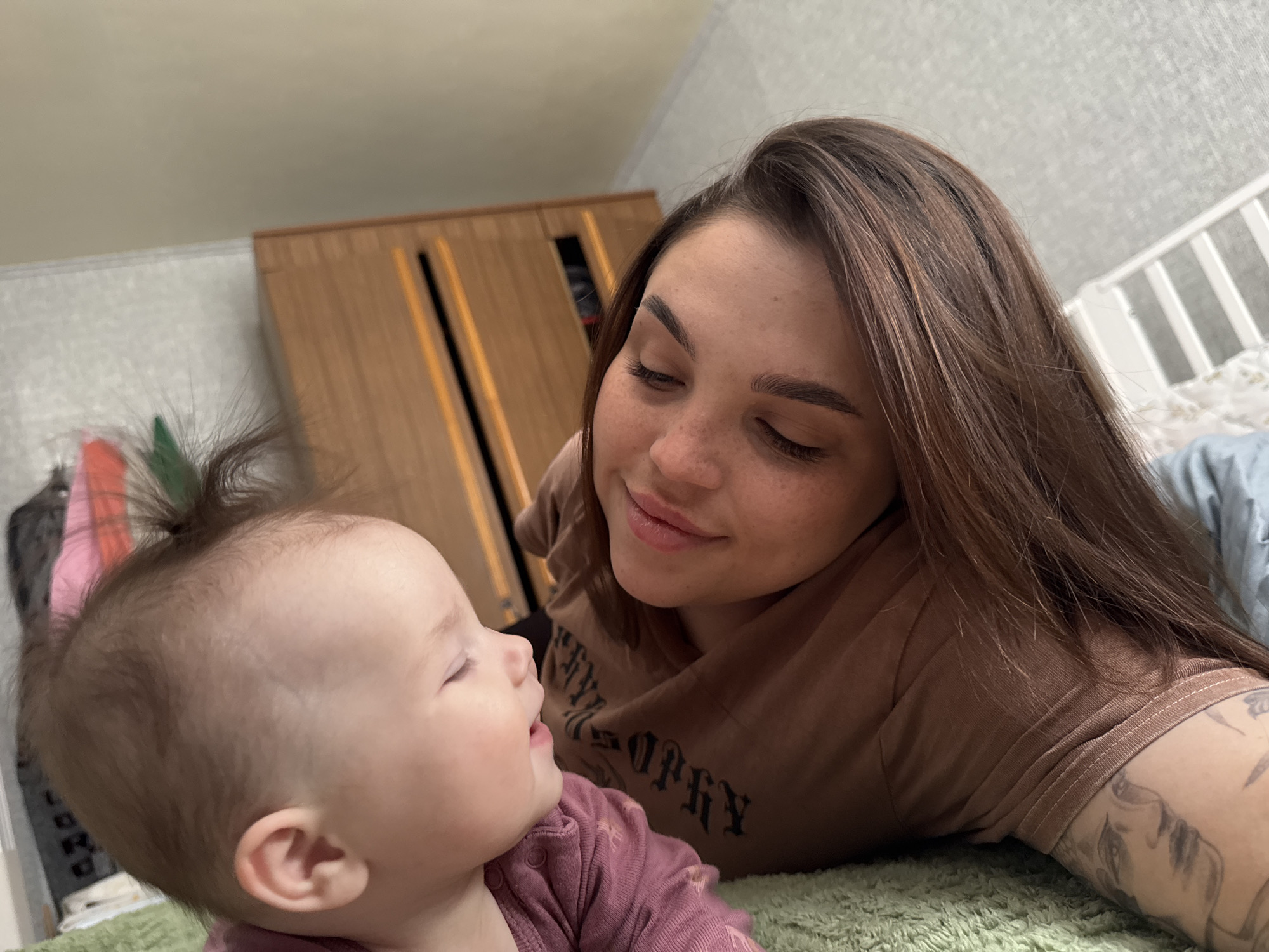 A medium, angled shot depicts a woman and a baby on a green, textured surface, likely a bed or floor covering, within an indoor setting. The woman, positioned on her side, gazes downward with a soft smile towards the baby. She has long, brown hair, fair skin, and freckles. Her dark eyebrows frame her eyes, which are directed downwards. She wears a brown t-shirt with dark, printed lettering and a tattoo is visible on her upper arm. The baby, in the foreground, is looking up towards the woman. The baby is wearing a pink long-sleeved garment and has fair skin and a tuft of hair tied on the top of their head. In the background, a light-colored wall and a wooden wardrobe are visible, indicating a domestic interior. The setting is most likely a residential building in Bataysk, Russia, based on the image's origin.