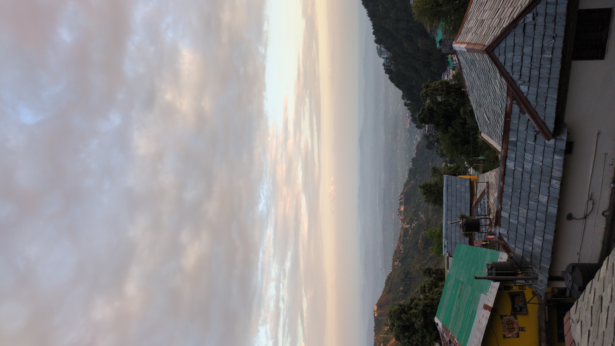 This image presents a serene mountain landscape viewed from an elevated perspective, likely at either sunrise or sunset. The sky dominates the upper portion of the frame, filled with diffused clouds that exhibit a gentle palette of light grey, pale blue, and soft yellow-orange, suggesting the atmospheric glow of twilight or dawn.
In the foreground, towards the right, are several roofs of buildings, possibly houses or guesthouses, built into the mountainside. Many of these roofs are constructed with dark grey, textured slate or stone tiles, showcasing traditional mountain architecture. One roof, lower down and to the left, is made of a contrasting bright green corrugated metal. Black water tanks and associated piping are visible on and around these structures. A yellow-painted wall below one of the tiled roofs features a framed piece of artwork or a decorative sign, although specific details are not discernible.
Beyond the immediate foreground, the landscape descends into a sprawling valley. This mid-ground and background feature layers of verdant hills and distant mountain ranges, with scattered settlements or towns visible across the slopes. A pervasive atmospheric haze lends a soft, muted blue-grey tone to the distant scenery, creating a sense of depth and tranquility. The setting evokes the peaceful and expansive beauty of a mountainous region, consistent with the provided location context of Haini, India. No individuals are present in the image, and no clear text is visible.