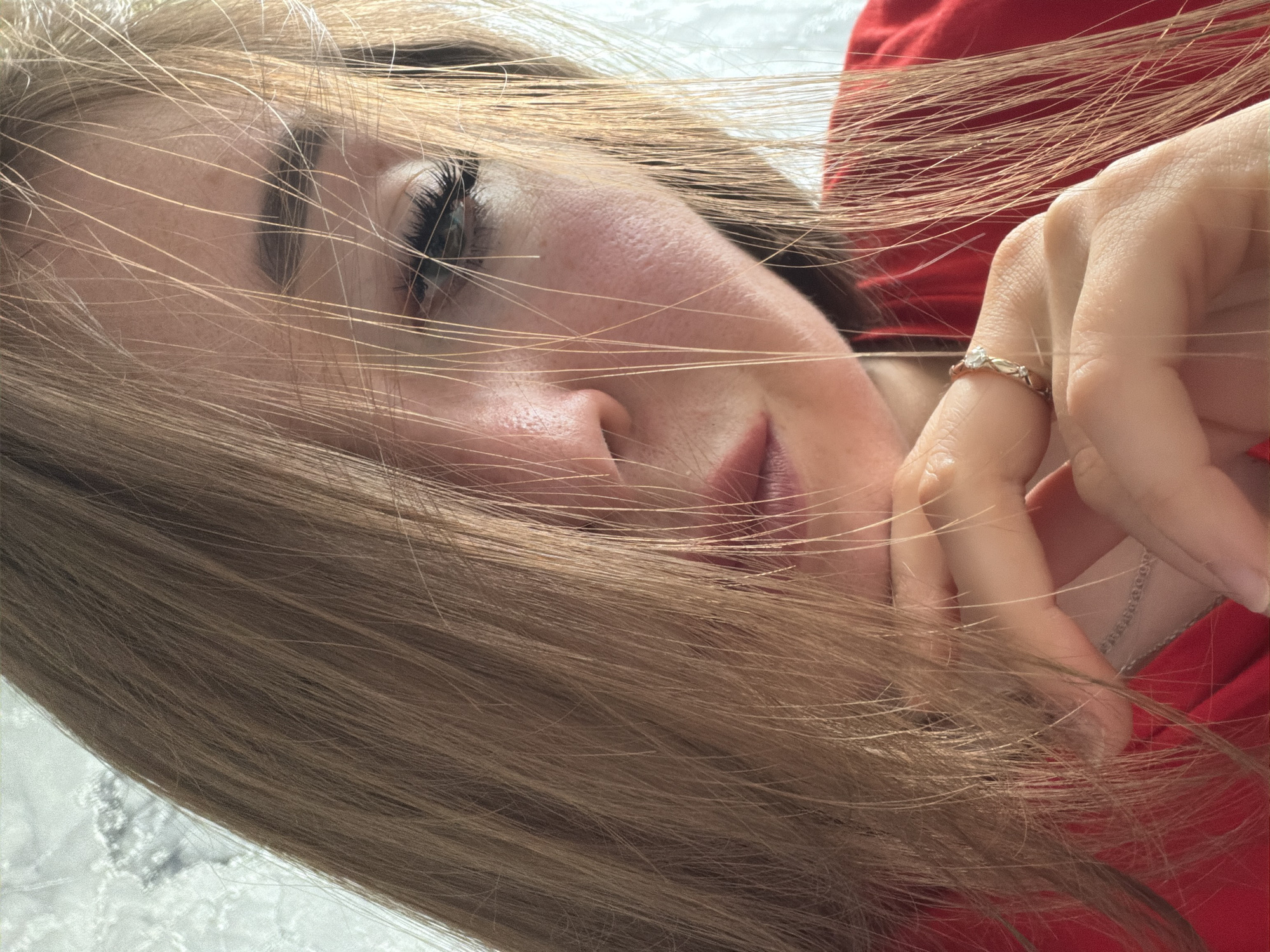A close-up, waist-up shot shows a young woman with long, light brown hair that partially obscures her face. Her head is tilted, and her chin rests on her hand, which is adorned with a ring. She is wearing a red top. The background appears to be a textured wall, possibly with a lace pattern. The lighting suggests an indoor setting. There is no information to determine the location as Старые Туймазы, Russia.