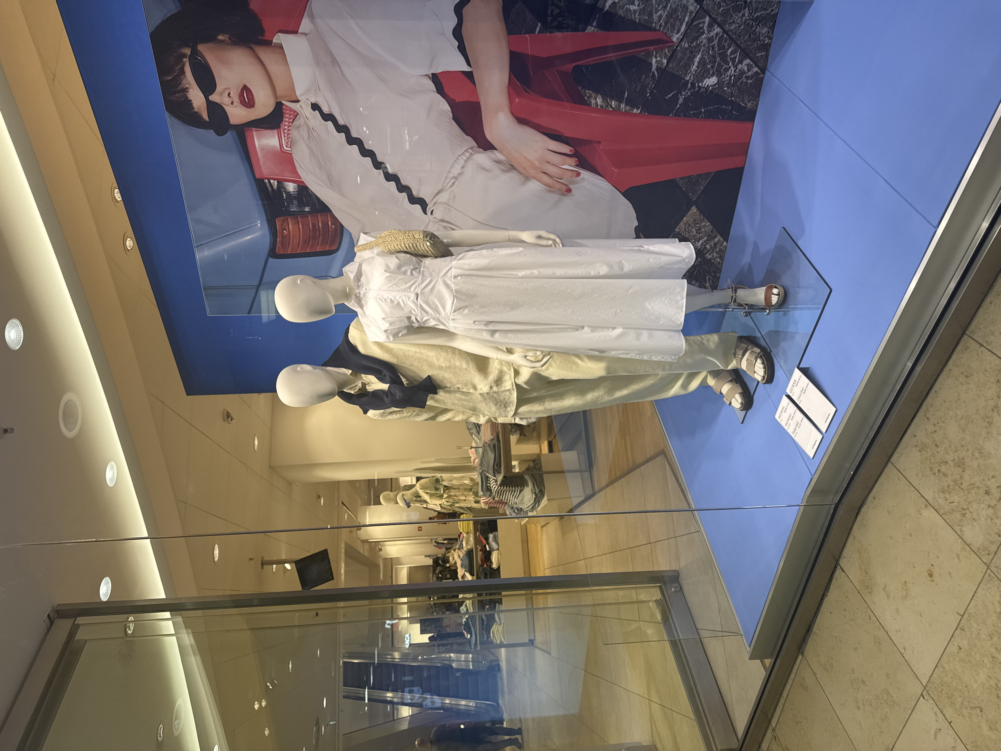 A store window display features two mannequins dressed in light-colored clothing. Above the mannequins, a large advertisement poster shows a woman wearing sunglasses and a white dress lying on a red chair. The mannequins are styled with sandals and a small straw purse on the top mannequin. The floor of the display is a solid blue color. The store window reflects the interior of a shopping mall, including escalators and other storefronts. Based on the presence of the brand names "MONCLER" and "GUESS" on cards in the window display, the store could be in an international shopping mall, with no specific visual evidence to confirm it's in Sofia, Bulgaria.
