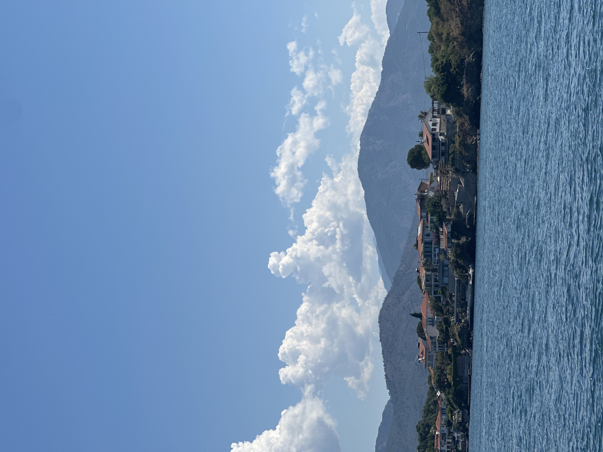 The image depicts a landscape of Fethiye, Turkey, featuring a clear blue sky with scattered cumulus clouds. Prominent mountains line the horizon, their slopes dotted with sparse vegetation. Along the coastline, numerous buildings, likely residential homes, are visible with orange-tiled roofs, nestled among trees. The foreground showcases a body of water, possibly the Mediterranean Sea or a bay, its surface textured with ripples. The image has been rotated, with the sky appearing on the left and the water on the right.