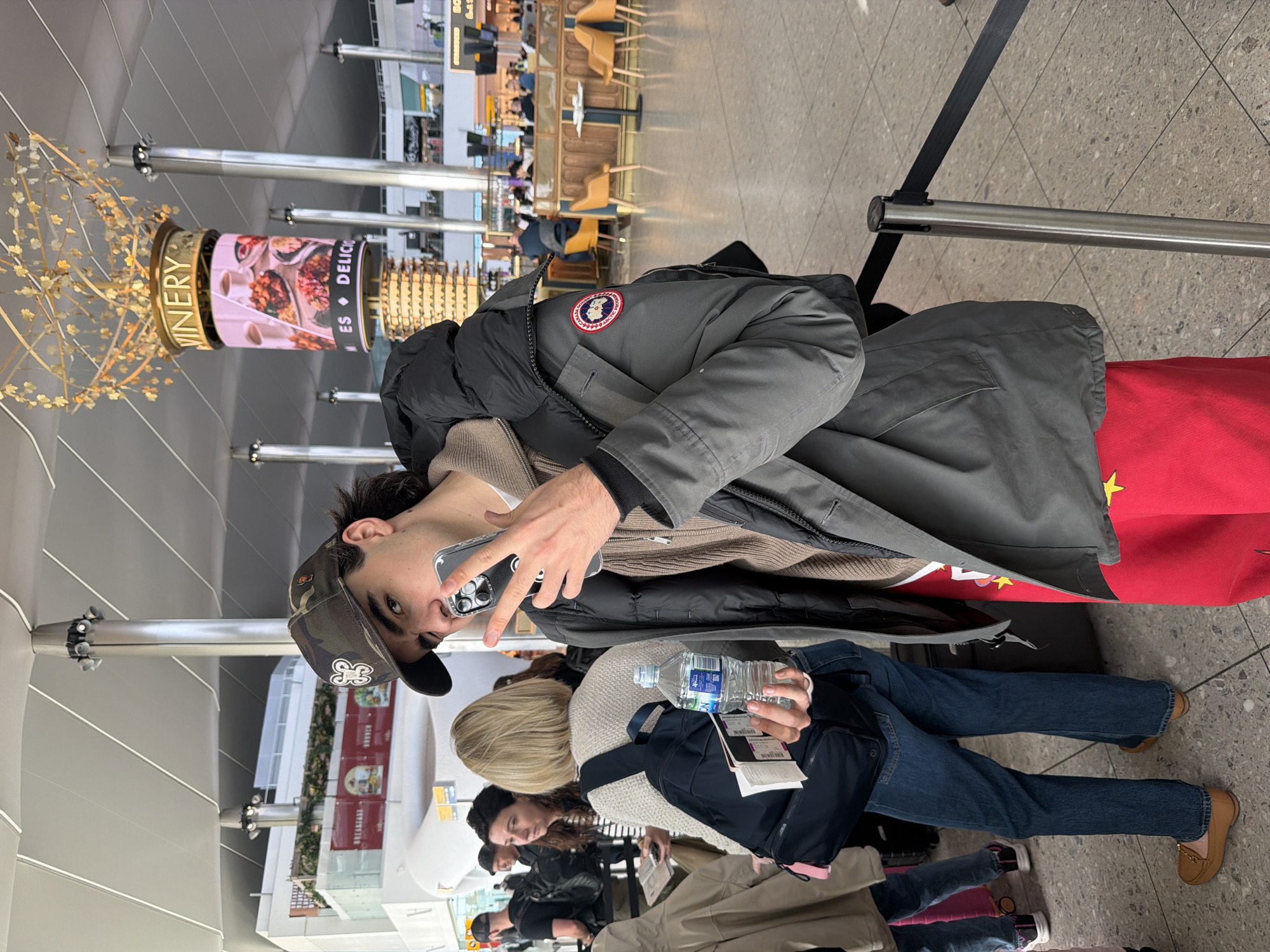 The image depicts a traveler in a grey Canada Goose jacket, red scarf, and camouflage baseball cap, holding a smartphone up to take a selfie in what appears to be an airport terminal. Other travelers are visible in the background. A sign reading "VINERY" is visible above. The location appears to be Heathrow Airport in London, United Kingdom.