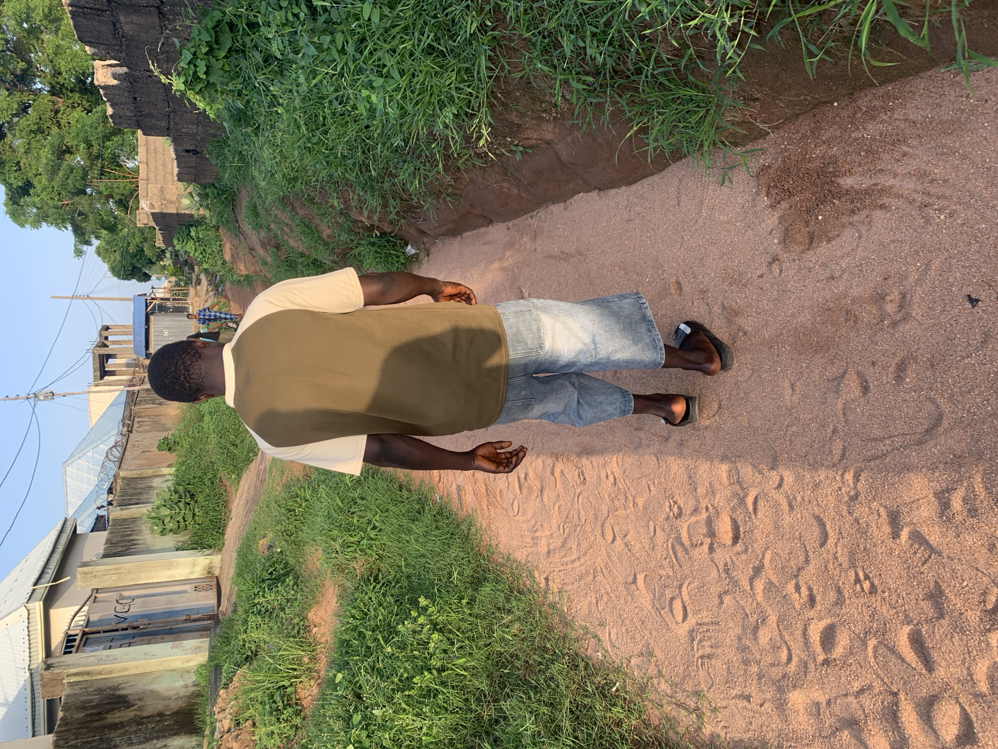 In Jalingo, Nigeria, a Black man walks away from the camera down a dusty, sandy path. He is wearing a short-sleeved shirt with a brown top and cream sleeves, and light-wash denim shorts. He has dark skin and short black hair. The path is flanked on the left by green grass and a wall with a gate, and on the right by a ditch filled with grass and a rough stone wall. In the background, buildings with metal roofs and barbed wire are visible, suggesting a residential or semi-urban area. The sky is clear and blue, indicating a sunny day. The overall impression is one of a simple, sun-drenched environment.