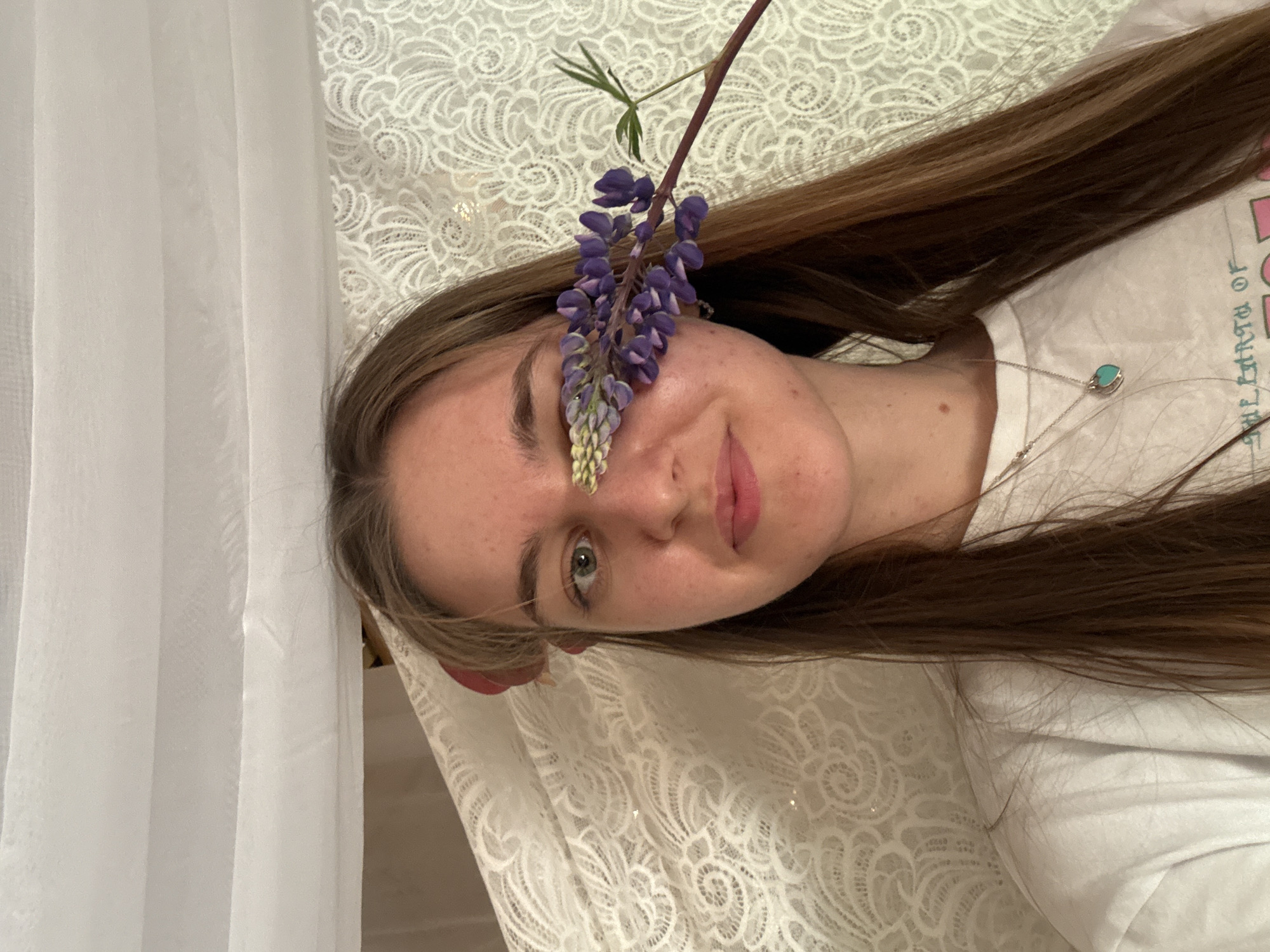 A woman lies on a white lace-patterned surface, possibly a bed or tablecloth, indoors. She faces the camera with a slight smile, and a purple lupine flower stalk is positioned over her left eye. She has light skin, brown hair, and is wearing a white t-shirt with writing on it and a necklace with a turquoise pendant. A sheer white curtain is visible to the left. There is no indication of Sibir', Russia.