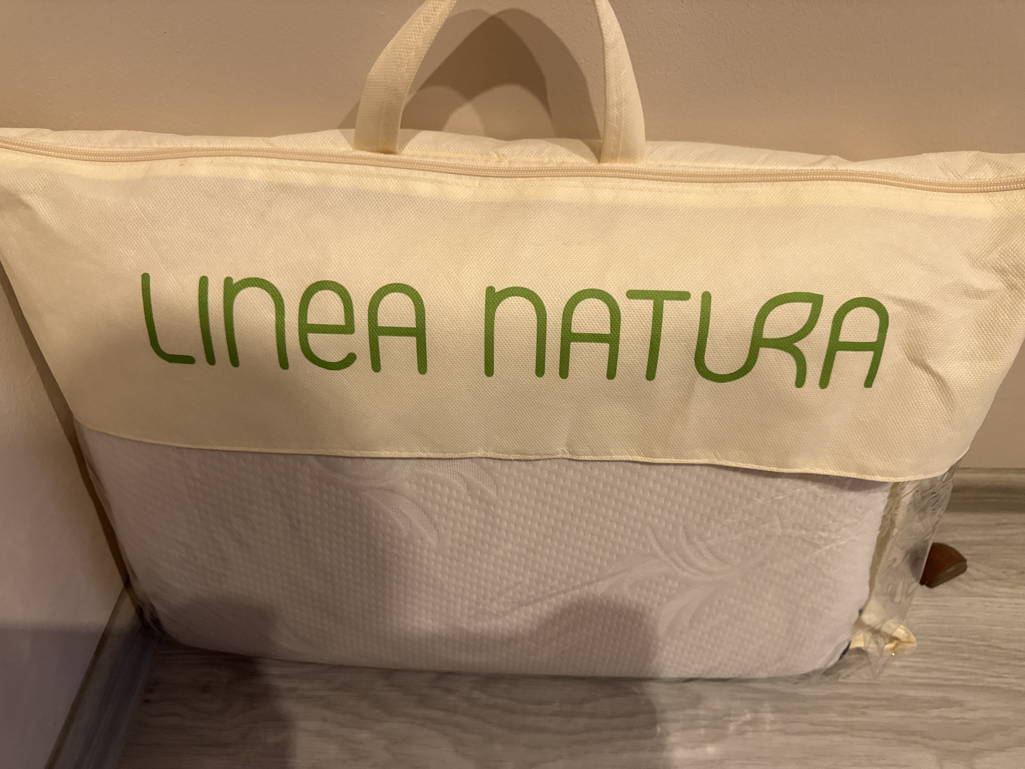 The image shows a pillow from Linea Natura in its carrying case, placed against a wall on a floor. The carrying case is a light beige color, with green lettering spelling out the brand name, and two beige handles at the top. The pillow inside the case appears to be white with a textured pattern. The floor is a light gray wood-like material. The location is unknown and cannot be confirmed to be in Lovech, Bulgaria based on the image alone.