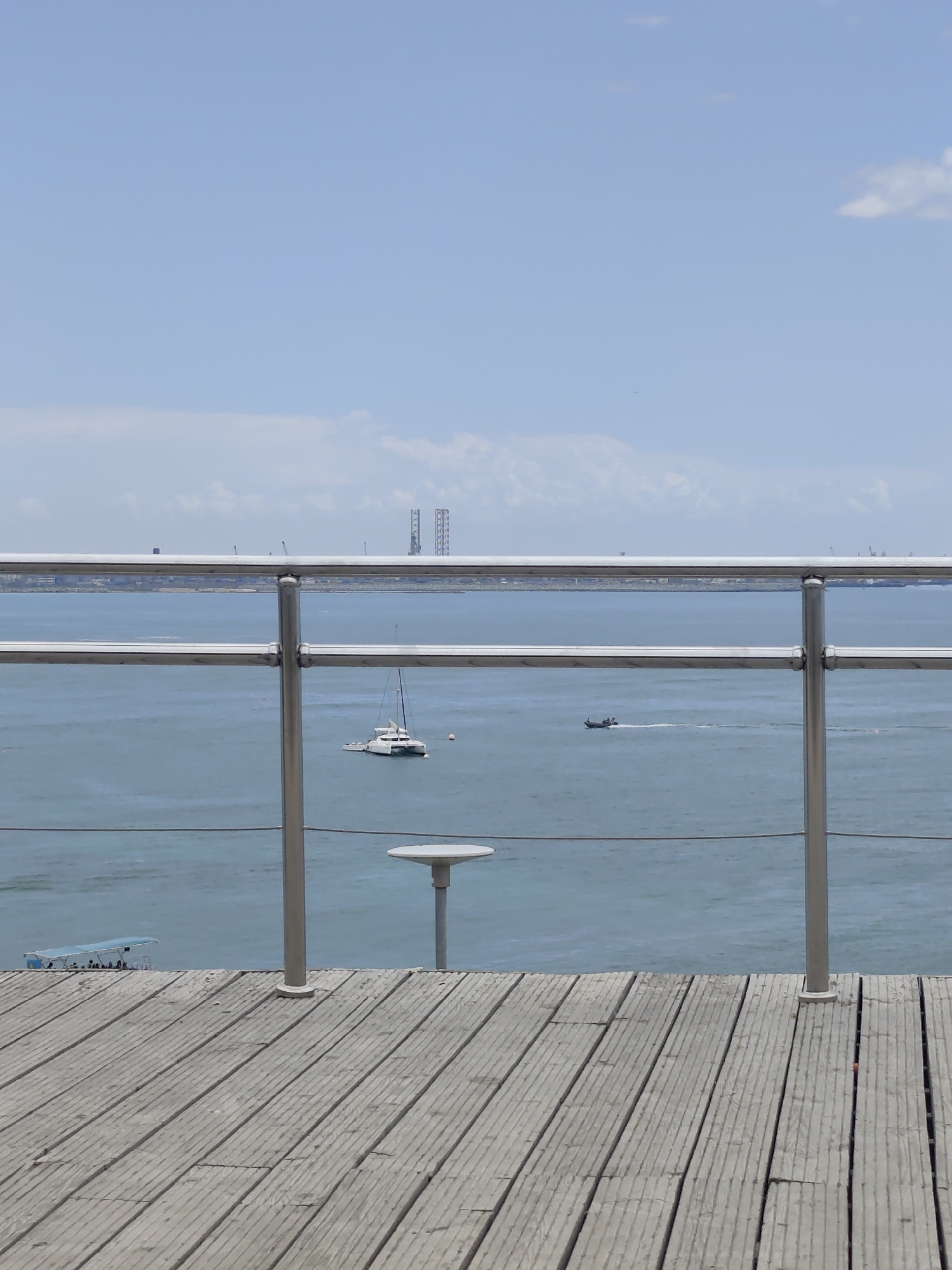 The image depicts a serene waterscape with a wooden deck in the foreground. A sleek, white catamaran with a black mast is anchored on the calm, blue water. Further in the distance, a smaller motorboat leaves a white wake as it speeds across the sea. The horizon is marked by a hazy industrial skyline with prominent lattice-like structures that resemble offshore oil rigs or cooling towers. A clear, light blue sky stretches above, with a few wispy clouds scattered across it. A metal railing with glass panels separates the deck from the water, offering an unobstructed view of the scene. On the deck, a small, round table is visible, suggesting a place for relaxation or dining. The overall atmosphere is tranquil and bright, indicative of a clear day. The presence of industrial structures on the horizon suggests a coastal location with a connection to maritime or industrial activities. No people are clearly visible. The time of day appears to be daytime, given the bright light. The weather seems pleasant and sunny. The location context provided is Aktau, Kazakhstan, which aligns with the presence of oil and gas infrastructure in coastal areas. There is no visible text in the image.