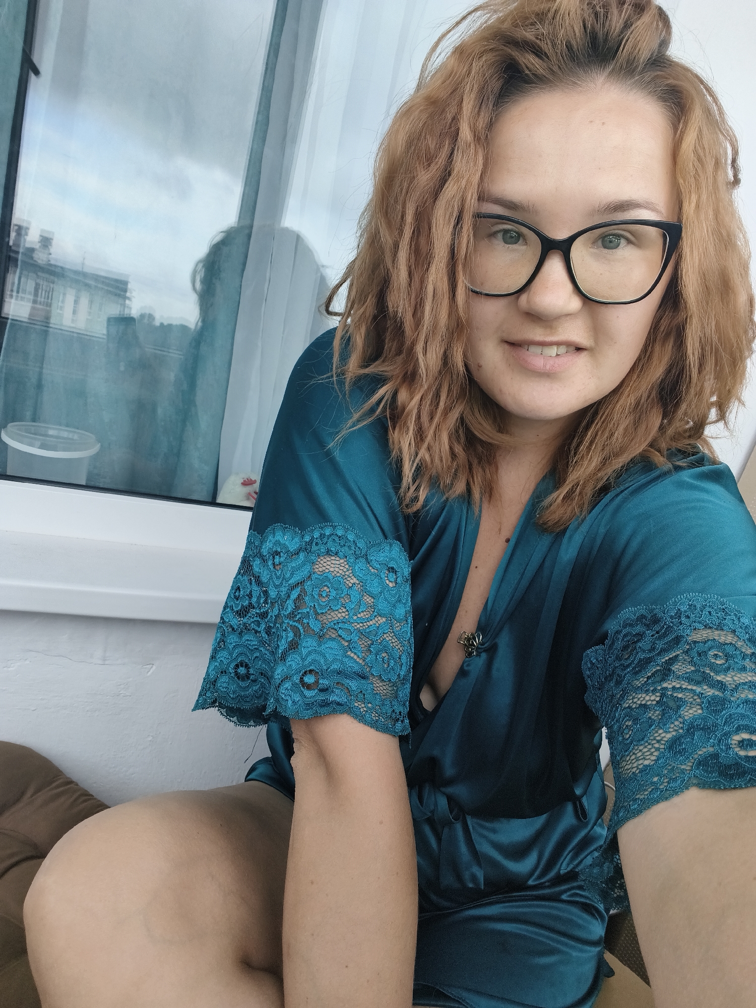 A woman with auburn hair and glasses sits on a balcony in Chaikovsky, Russia. She is wearing a teal silk robe with lace trim. Her left arm is resting on her leg, and she is looking at the camera. The background shows a window with a reflection of buildings and a cloudy sky.