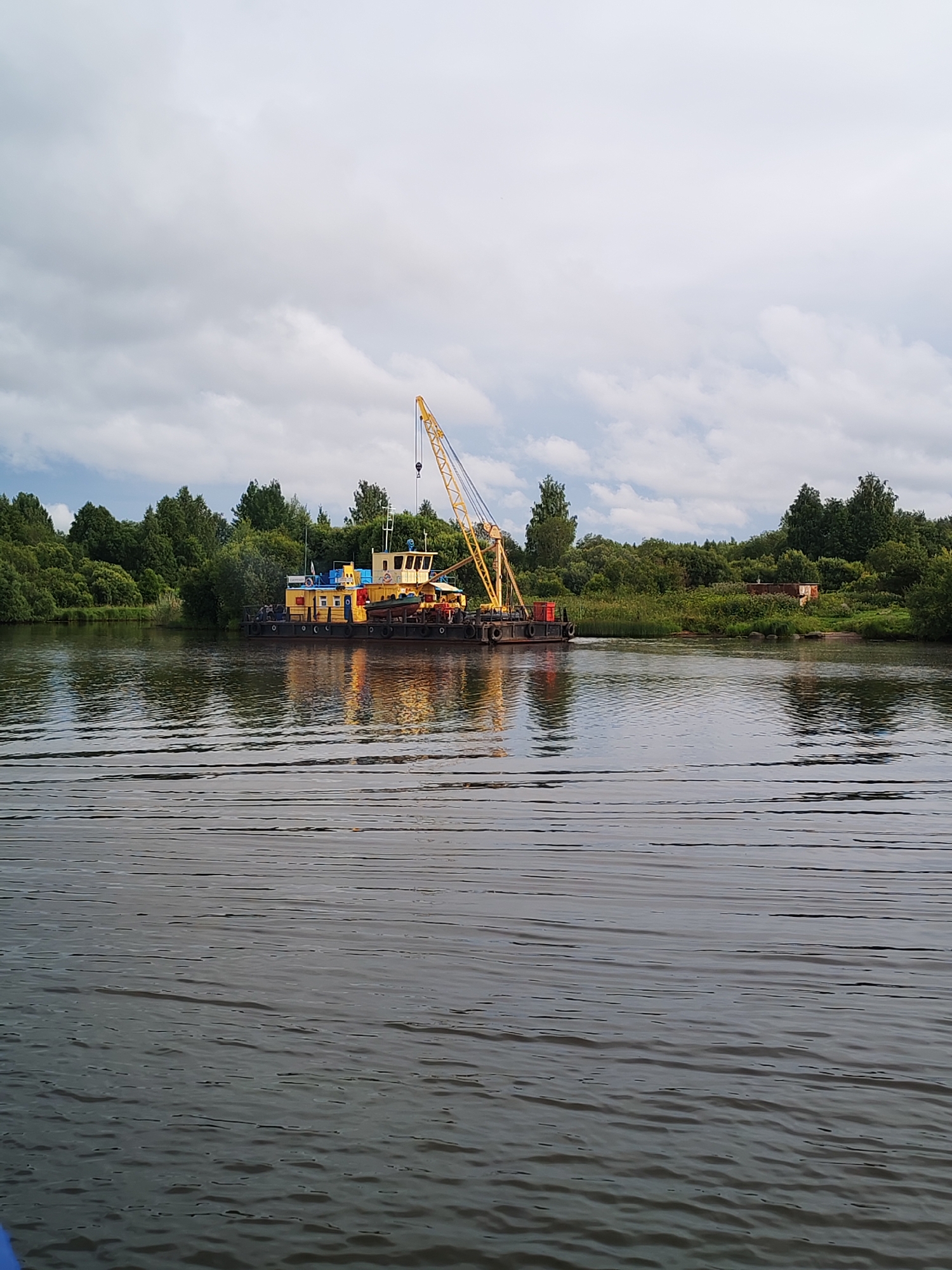 The image shows a crane barge on a body of water, likely a river, with a densely wooded shoreline in the background. The barge is yellow and black, with a tall yellow crane mounted on it. The barge appears to be stationary. The water is rippled, suggesting some movement. The sky is overcast with clouds. The general impression is one of an industrial or work barge on a natural waterway. The location is identified as Kargulino, Russia.