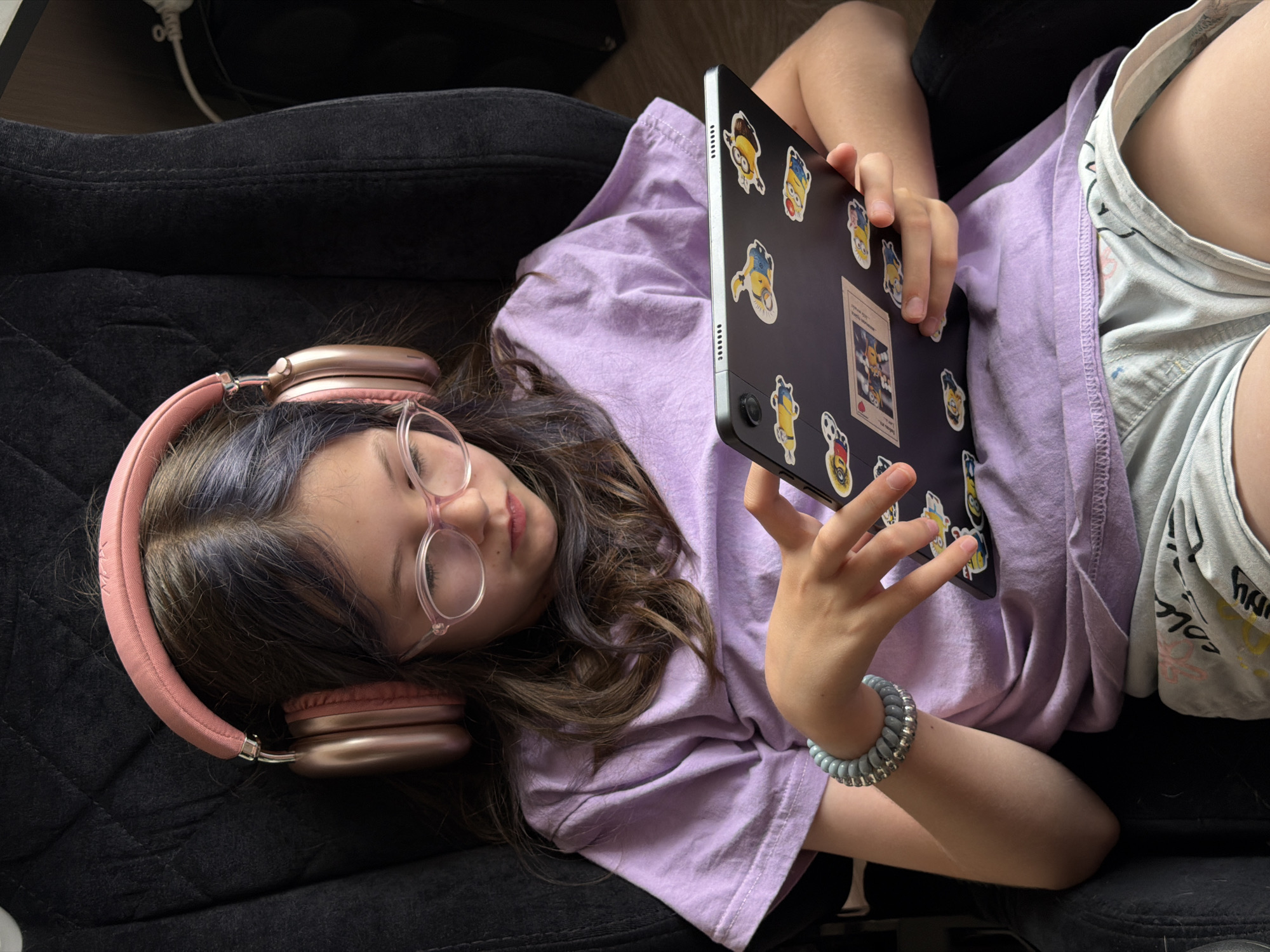 A young girl with long brown hair and glasses is sitting in a black chair, wearing pink headphones, a light purple t-shirt, and light blue shorts. She is holding a black tablet decorated with Minion stickers and is looking at the screen, her fingers touching it. The background is dark and appears to be indoors. The image does not contain any information that places it in Birobidzhan, Russia.