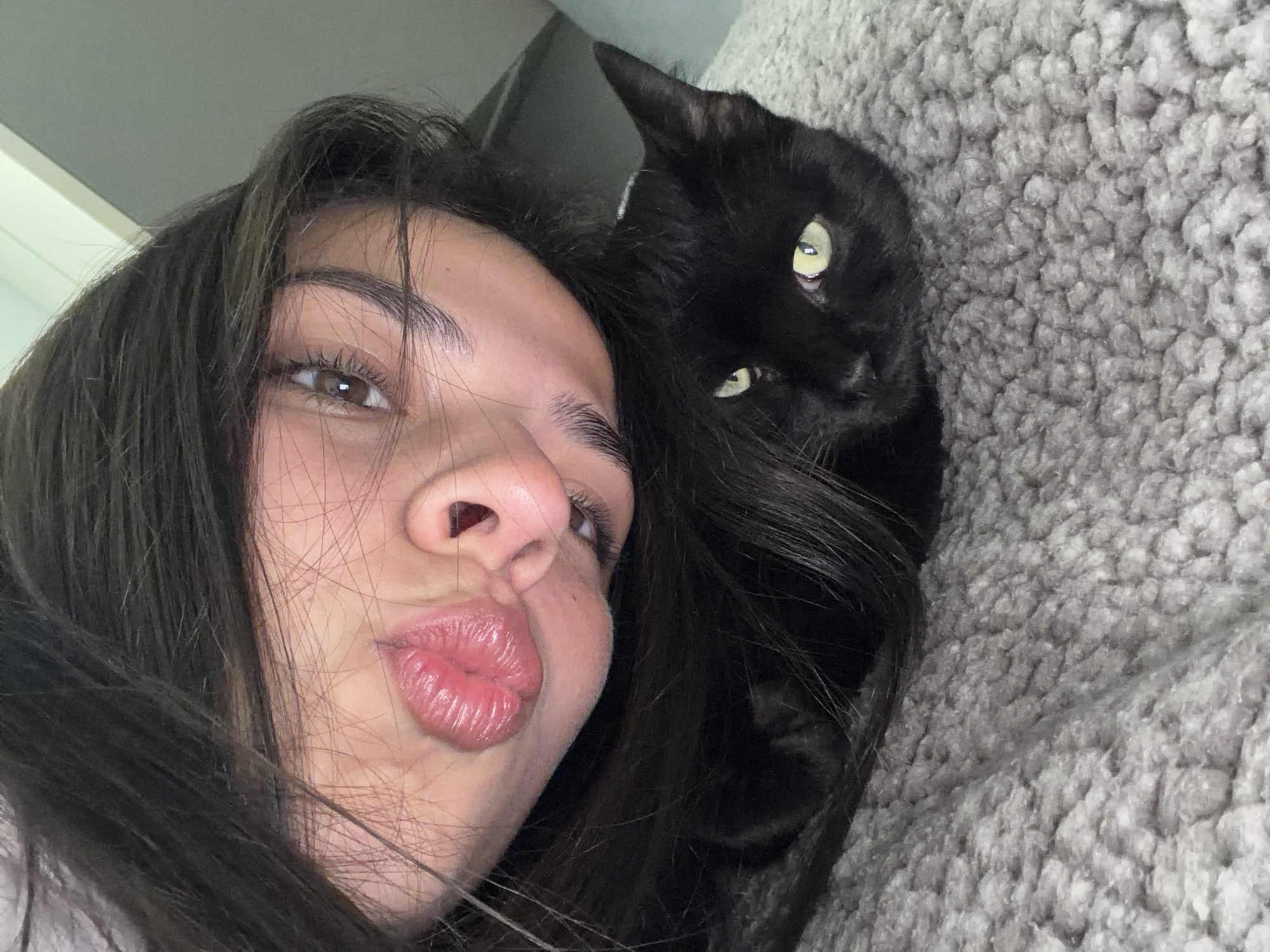 A young woman with dark hair is taking a selfie with a black cat on a gray, textured surface, likely a blanket or bedspread. The woman is in the foreground, her face angled towards the camera with her lips pursed in a kissing expression. The cat is positioned behind her head and appears to be looking at the camera with bright green eyes. There is no indication of a location in Gabrovo, Bulgaria.