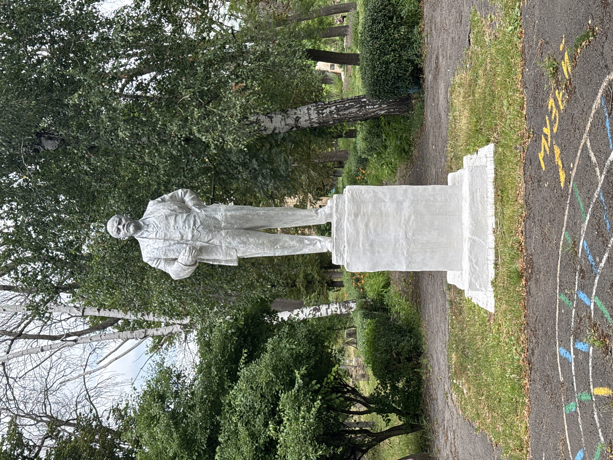 The image depicts a full-body, light-colored statue of Vladimir Lenin standing on a white pedestal in a park setting. Lenin is depicted in a suit, with his hands clasped in front of him. The statue is surrounded by lush green trees and bushes, with a paved walkway leading towards it. In the foreground, a section of asphalt with chalk markings for a game, possibly hopscotch, is visible. The presence of a statue of Lenin and the chalk markings suggest this location is likely a public space in a city or town. Given the context provided, this scene is located in Seleznivka, Ukraine. The overall atmosphere is one of a quiet, possibly slightly overgrown, public area with a prominent historical monument.