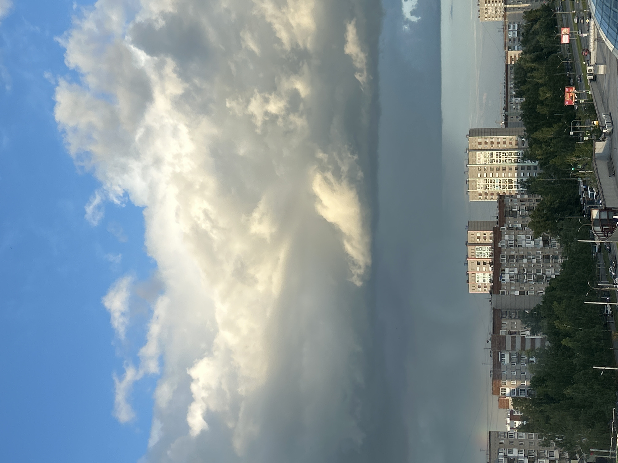 The image shows a view of the city of Izhevsk, Russia. On the left side of the frame, a blue sky with scattered white clouds transitions into a massive, dark storm cloud that dominates the center of the image. The right side of the frame captures a densely populated urban landscape, with rows of tall apartment buildings, trees, roads with vehicles, and streetlights. The city appears to be juxtaposed against the approaching storm, creating a sharp contrast between the sunny skies and the impending darkness.