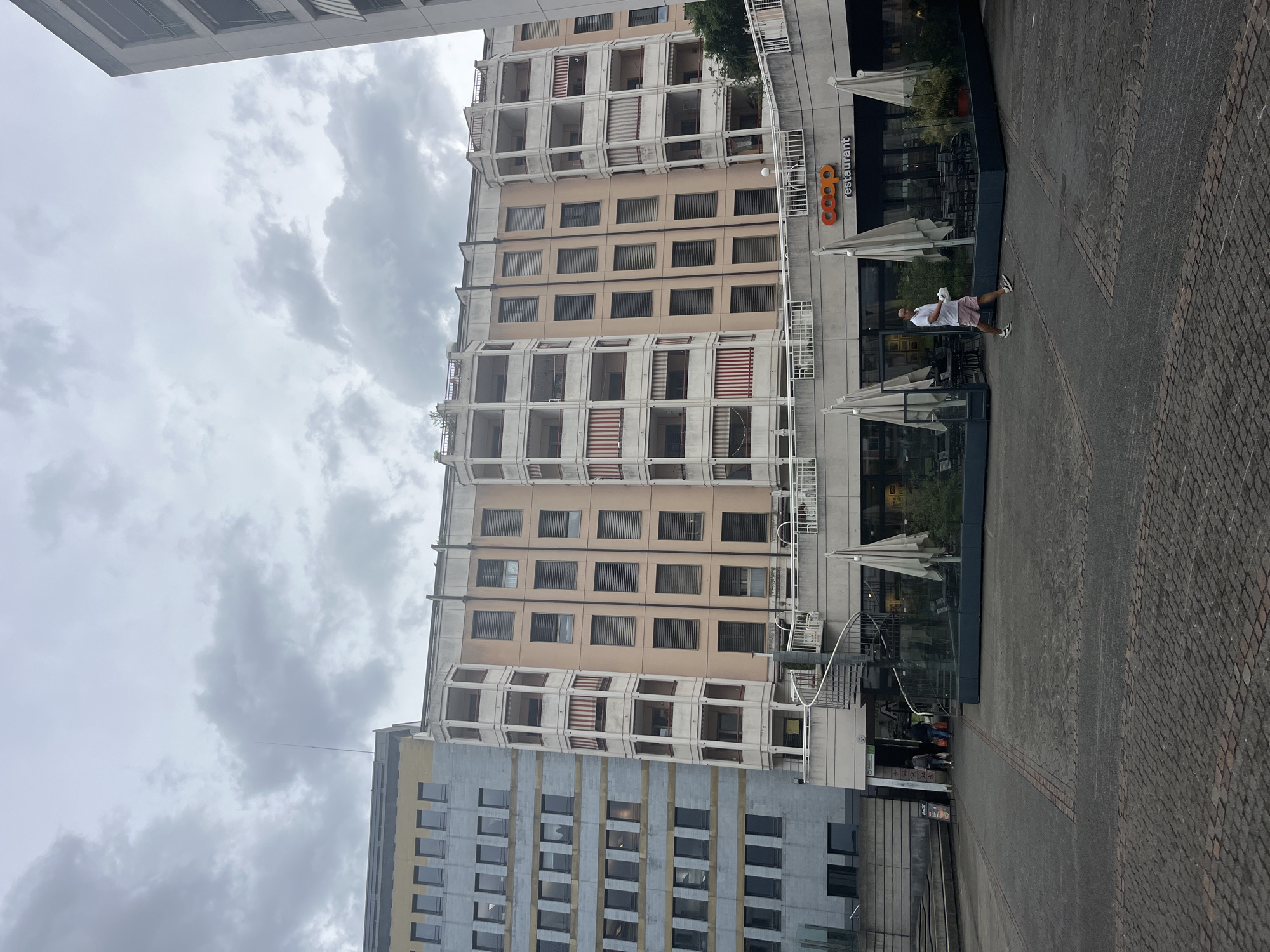 The image depicts a street scene in front of a building in Geneva, Switzerland. The building is multi-story with a light peach-colored facade and numerous balconies with white railings. To the left of this building, another multi-story building with a light blue and grey facade is visible. Both buildings appear to be residential or office spaces.
At the base of the peach-colored building, there is a Coop restaurant with large glass windows and outdoor seating. Several white umbrellas are set up for shade. A spiral staircase is attached to the side of the building, leading up to the upper levels.
In the foreground, a paved public square is visible, made of grey cobblestones and brick patterns. A man is walking across the square from right to left, wearing a white polo shirt and pink shorts, carrying a white object in his right hand. Two other individuals are walking away from the camera towards the entrance of the building. The sky is overcast with grey clouds, suggesting a cloudy day.