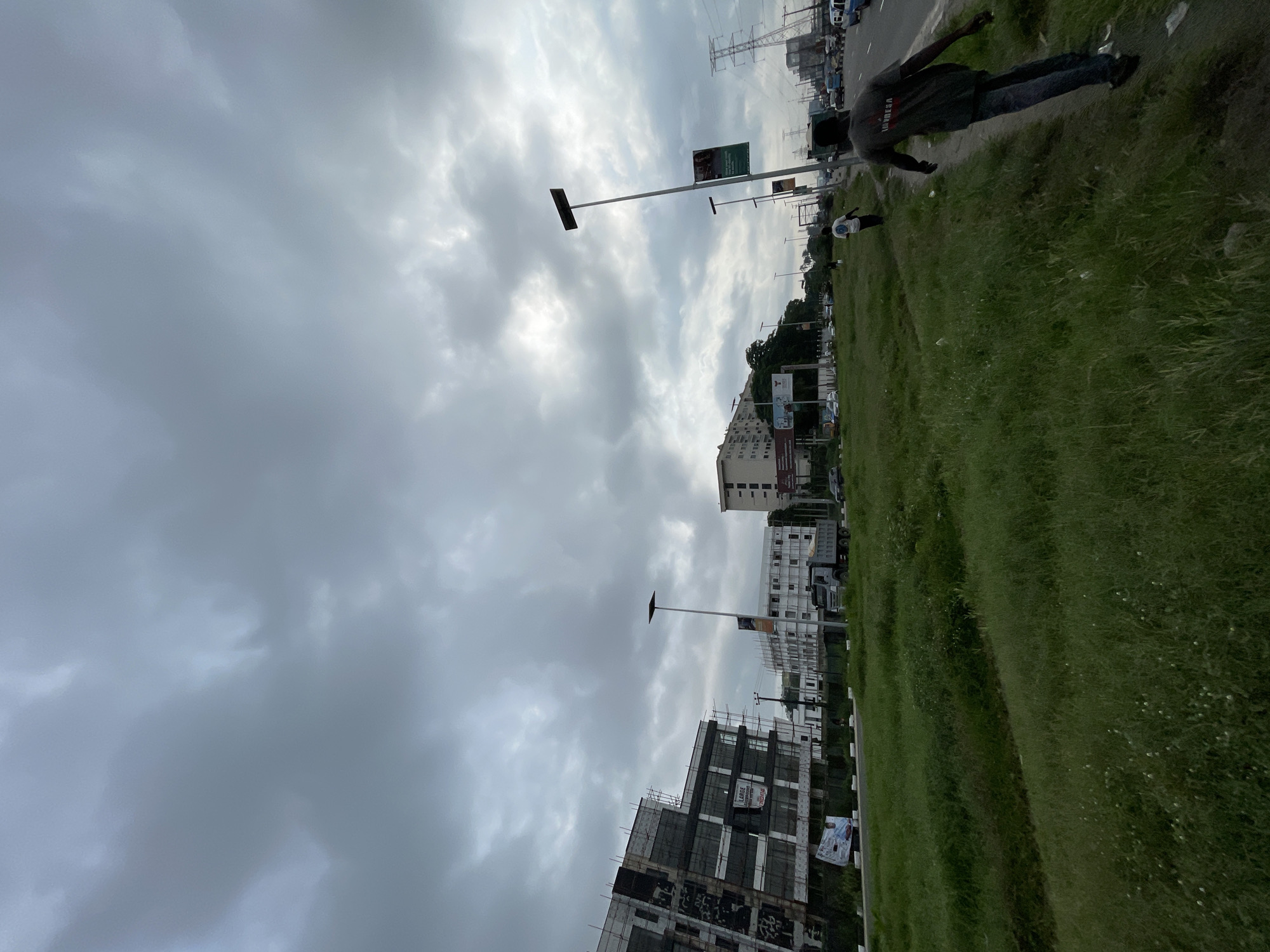 In Igboefon, Nigeria, under a cloudy sky, a street scene unfolds with an unfinished building on the left, its facade adorned with scaffolding and construction materials. Adjacent to it, another building stands complete, its windows reflecting the overcast sky. Across the road, a truck is parked. The foreground is dominated by a grassy median, bordered by a sidewalk where two individuals walk. One person, wearing a dark t-shirt and jeans, walks away from the camera, while another, in a white t-shirt and shorts, walks further ahead. The road is busy with vehicles, suggesting an active urban environment. Billboards and streetlights punctuate the landscape, contributing to the city's dynamic character. A power pylon is visible in the distance, hinting at the city's infrastructure.