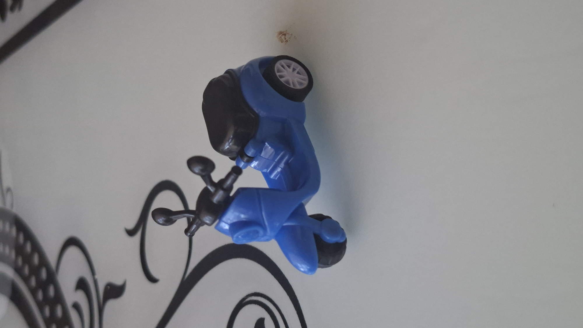 A small, blue toy scooter with a black seat and black handlebars is positioned on a light-colored, flat surface. The scooter is detailed with two wheels at the rear and one at the front, with a light-colored rim on the rear wheel. The background shows a portion of a decorative pattern with black swirls and dots on a white surface. There is a small, light brown smudge to the right of the scooter. There are no people, actions, or interactions visible. The scene is devoid of any contextual information that would allow for the determination of its location in Voinova, Moldova.