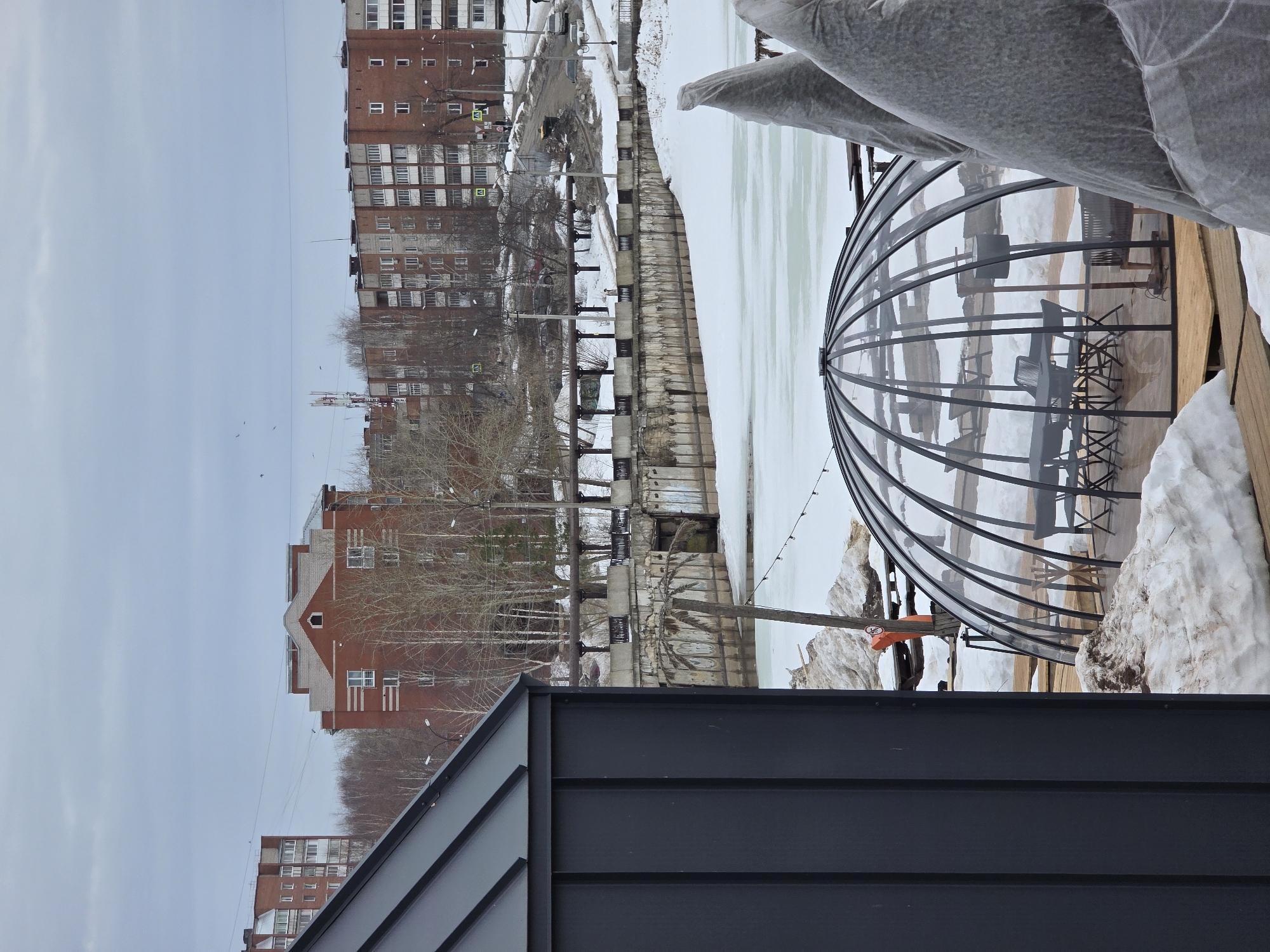 This is a view over the top of a dark grey or black roof, with a river or canal in the distance. The canal is frozen over, and there are buildings across the canal, including apartment blocks and a red brick building. On the right of the image is a transparent dome structure, possibly for outdoor seating. There is snow visible around the dome. Part of a covered object is also in the shot. The photo was taken in the city of Votkinsk, Russia.