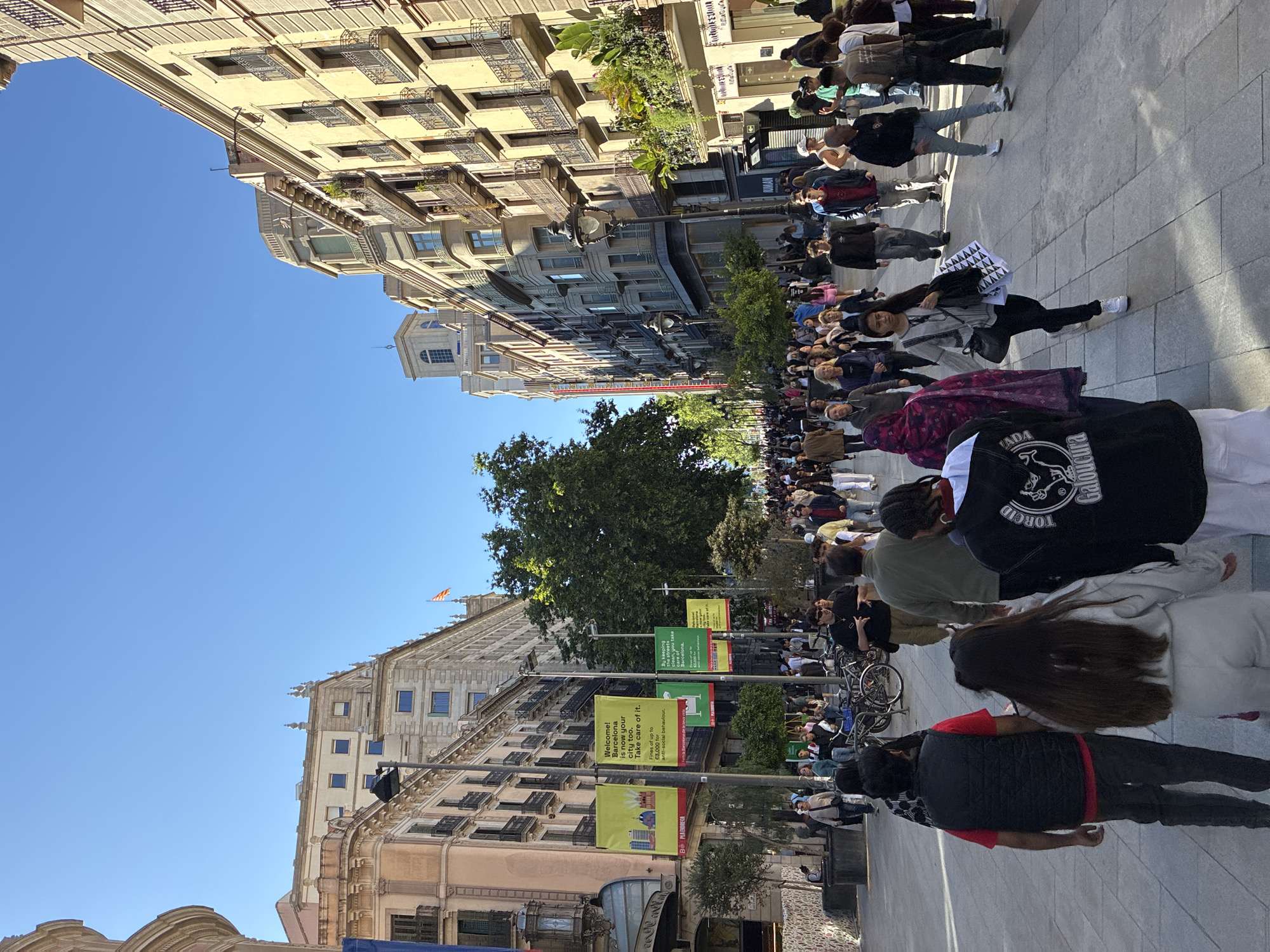 The image depicts a pedestrian street scene in Barcelona, Spain, populated by a crowd of people. The buildings lining the street exhibit classical European architecture. Several banners hang along the street with messages related to public behavior and cleanliness, mentioning fines for antisocial conduct. A Catalan flag is visible atop one of the buildings. The sky is clear and blue.