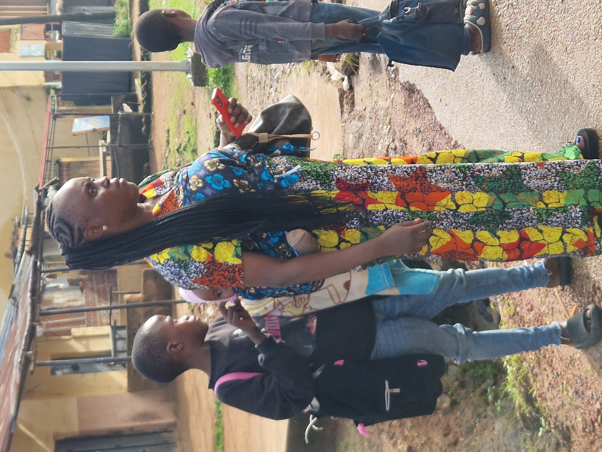 A full shot captures a group of people, likely a family, standing outdoors in what appears to be an urban area, possibly in New Stadium, Nigeria. On the right, a woman in a vibrant, multi-colored dress is holding a red smartphone. She has her hair braided and is wearing a gold earring. She is also carrying a dark bag. In the foreground and slightly to the left, a young boy in a black sweatshirt and blue jeans is facing left, wearing a black backpack with pink accents. To his right, a smaller child, possibly a baby, is being held by the woman, wrapped in a light-colored fabric with blue and yellow designs. Further to the right and slightly behind the woman, another young boy in a grey hooded sweatshirt and blue jeans stands facing away from the camera. He is carrying a blue bag. The ground is a mix of dirt and gravel, with some patches of grass. In the background, there are buildings with corrugated metal roofs, suggesting a semi-developed or informal settlement. The overall lighting indicates it is daytime, with some shadows suggesting the sun is present.