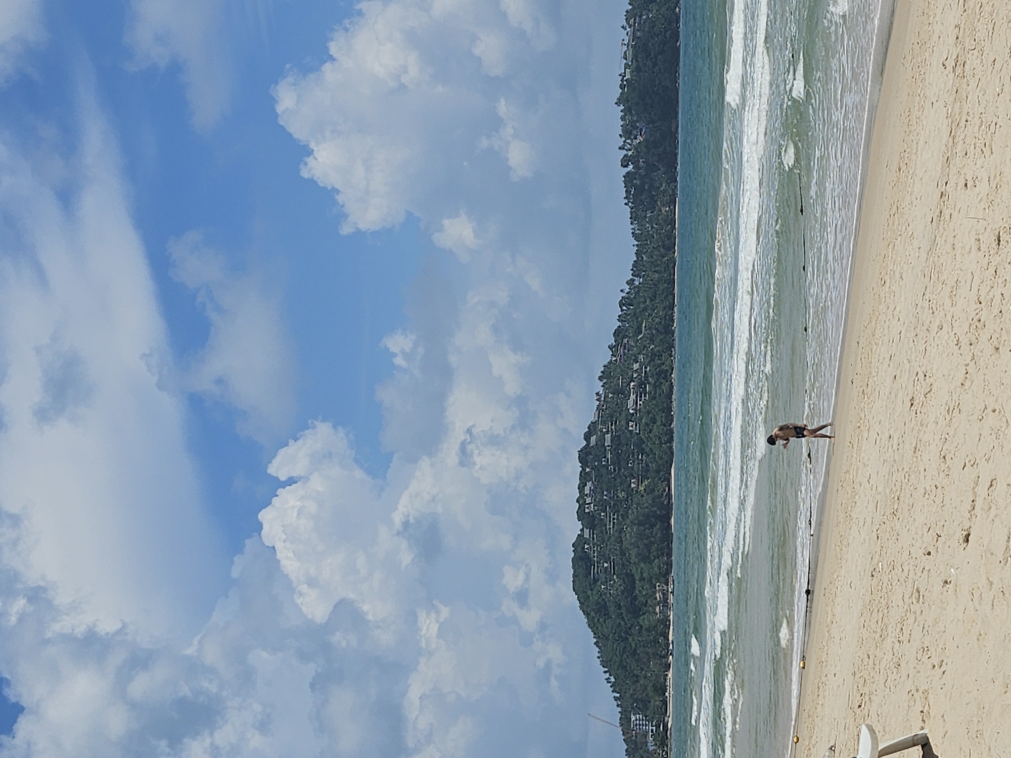 A lone male is walking along the shoreline of a sandy beach in Choeng Thale, Thailand. He is wearing dark shorts and appears to be looking down at the sand. The ocean has moderate waves, with white crests indicating movement. In the distance, a lush green hill is visible, dotted with white buildings that suggest a resort or residential area. The sky is a vibrant blue, with scattered white cumulus clouds. The sand is a light tan color, with some scattered debris and footprints visible. A white beach chair is partially visible in the lower left corner of the frame.