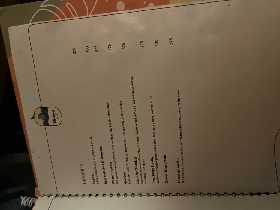 The image displays an open, spiral-bound dessert menu from "Windsong CAFE," with its logo visible at the top left. The page is titled "DESSERTS" and lists several items with descriptions and prices.
The desserts featured are:
* Tiramisu, described as offering "unique flavours of coffee and cream," priced at 250.
* New York Style Cheesecake, also priced at 250.
* Walnut Brownie, described as "Our signature walnut brownie with ice-cream and hot chocolate sauce," priced at 325.
* Gud Bud, an ice-cream sundae described as for those who "Love ice-creams & toppings, then dig into this specially curated sundae," priced at 275.
* Death by Chocolate, featuring "chocolate brownie, white chocolate chips, wafers served with a sizzling hot sauce on top, served with ice-cream," priced at 325.
* Oreo fudge Sunday, a "Vanilla ice-cream topped with hot chocolate sauce / salted caramel sauce," priced at 275.
* Choice Of Ice Cream, priced at 120.
* Chocolate Fondue, described as "Our signature chocolate fondue, with seasonal fruits and cakes, for the table," priced at 275.
The background shows a blurred surface with light orange and brown organic patterns, suggesting a table in an indoor setting. The lighting appears to be artificial. The image is captured at an angle, with the menu slightly rotated.