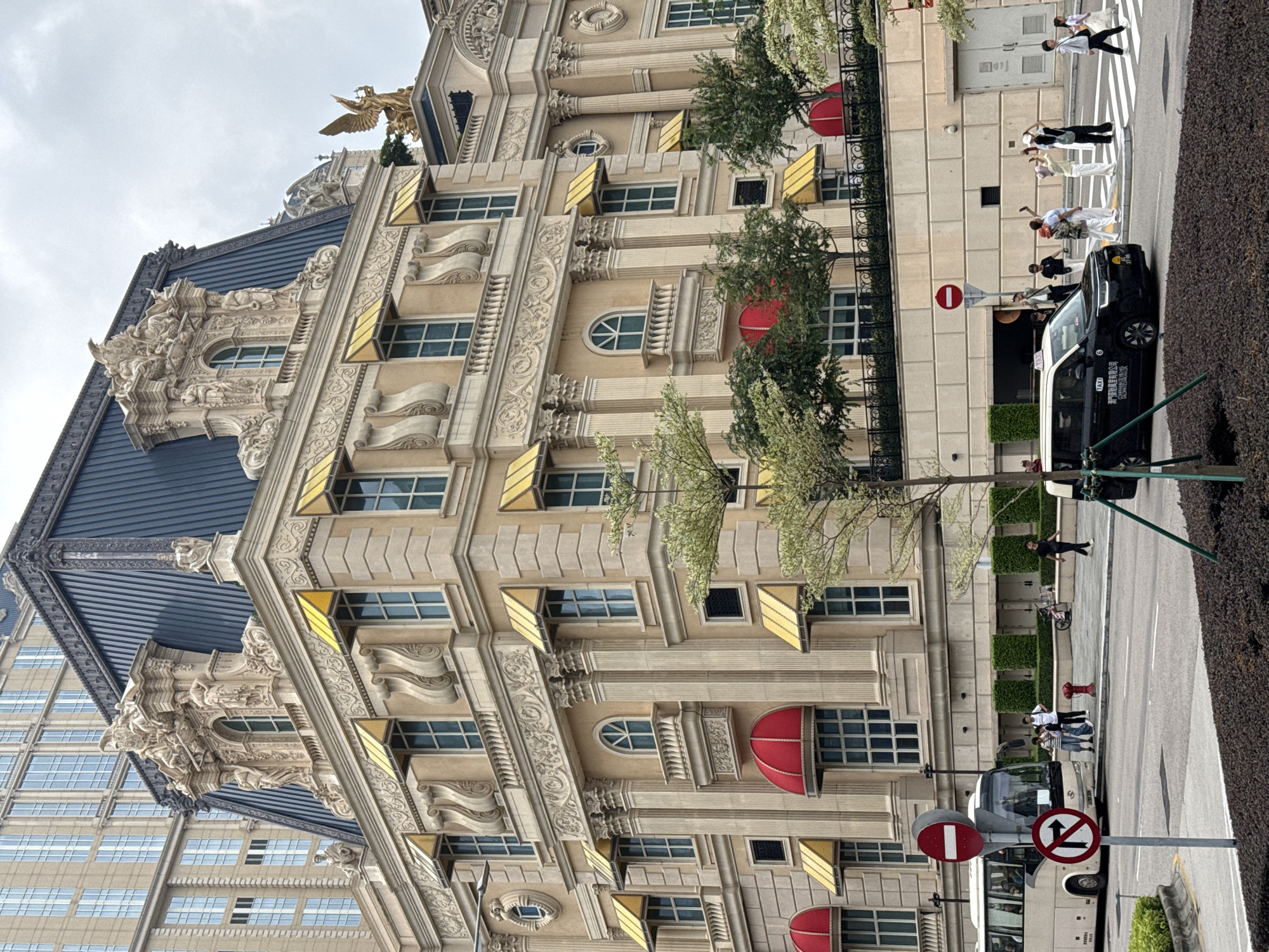 The image captures a street scene in Unknown, Macao, showcasing a blend of architectural styles and urban activity. The primary focus is a multi-story building with ornate detailing, including gold-colored awnings and classical-style embellishments. A dark blue roof with intricate carvings is also visible. The building is situated near a street, where a black taxi is parked alongside a crosswalk. Several pedestrians are crossing the street, while others are standing on the sidewalk. A bus is also present. The environment is urban, with street signs and greenery visible.
