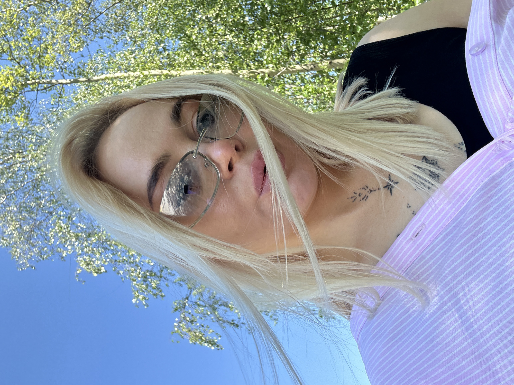An eye-level outdoor portrait features a young woman with long, straight blonde hair, looking directly at the camera. She wears round, wire-rimmed sunglasses that reflect the bright sky and tree canopy. Her lips are slightly parted, and her tongue is visible. She is dressed in a black top with a light purple and white striped button-up shirt worn over it, partially unbuttoned.
The image is captured from a low angle, pointing upwards towards a clear blue sky. Lush green tree foliage with small leaves is visible in the background, suggesting a sunny day in spring or early summer. Several small, dark tattoos are visible on her left shoulder and neck area, including a star-like design and other abstract patterns. The lighting indicates daytime with bright sunshine. There is no visible text in the image. The overall impression is a casual, relaxed selfie taken outdoors.