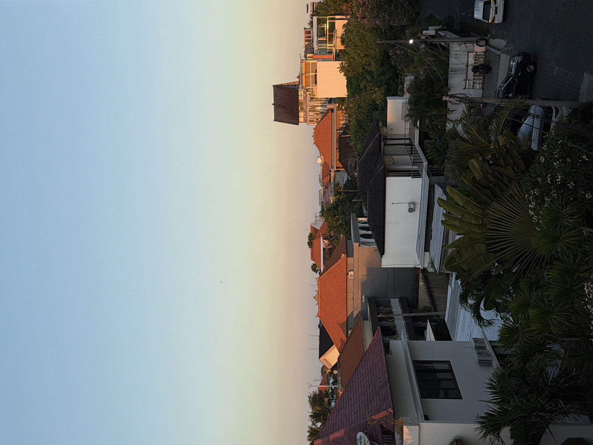 The image depicts an aerial view of a residential area in North Kuta, Indonesia, during either sunrise or sunset. The sky is a gradient of pale blue transitioning to a soft yellow or orange hue near the horizon. Numerous rooftops of varying architectural styles are visible, with prominent red-tiled roofs and some with darker, flat roofs. Buildings range from multi-story structures to smaller homes, some featuring balconies and air conditioning units. Lush green foliage, including palm trees and flowering bushes, is interspersed among the buildings, suggesting a tropical climate. A paved road is visible on the right side of the image with two vehicles parked or driving, one white SUV and a dark sedan with a roof rack. A motorcycle is also parked near a wall. The overall impression is a peaceful urban landscape bathed in the warm, soft light of dawn or dusk.