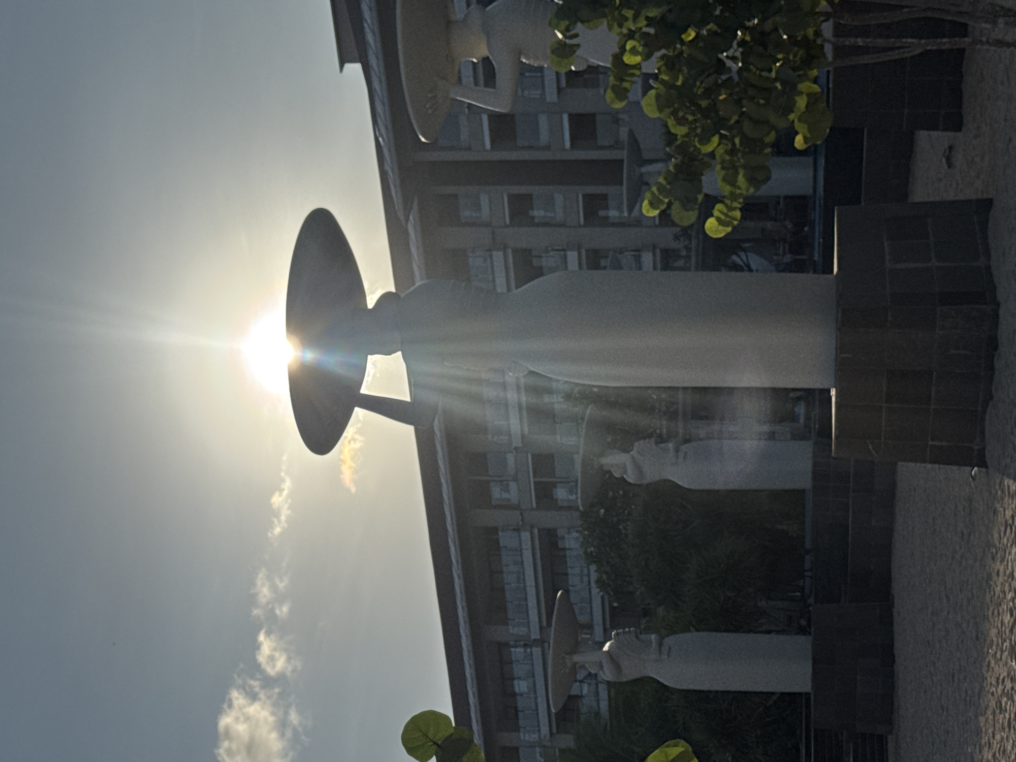 This outdoor shot shows a row of statues with dish-shaped hats in Kuta Selatan, Indonesia. The statues are white and stand on dark, square pedestals. The sun is directly behind one of the statues, creating a strong glare. A building with many windows is behind the statues. Some greenery is visible in the foreground and around the statues. The sky is blue with some clouds. The ground appears to be a light-colored, coarse material.