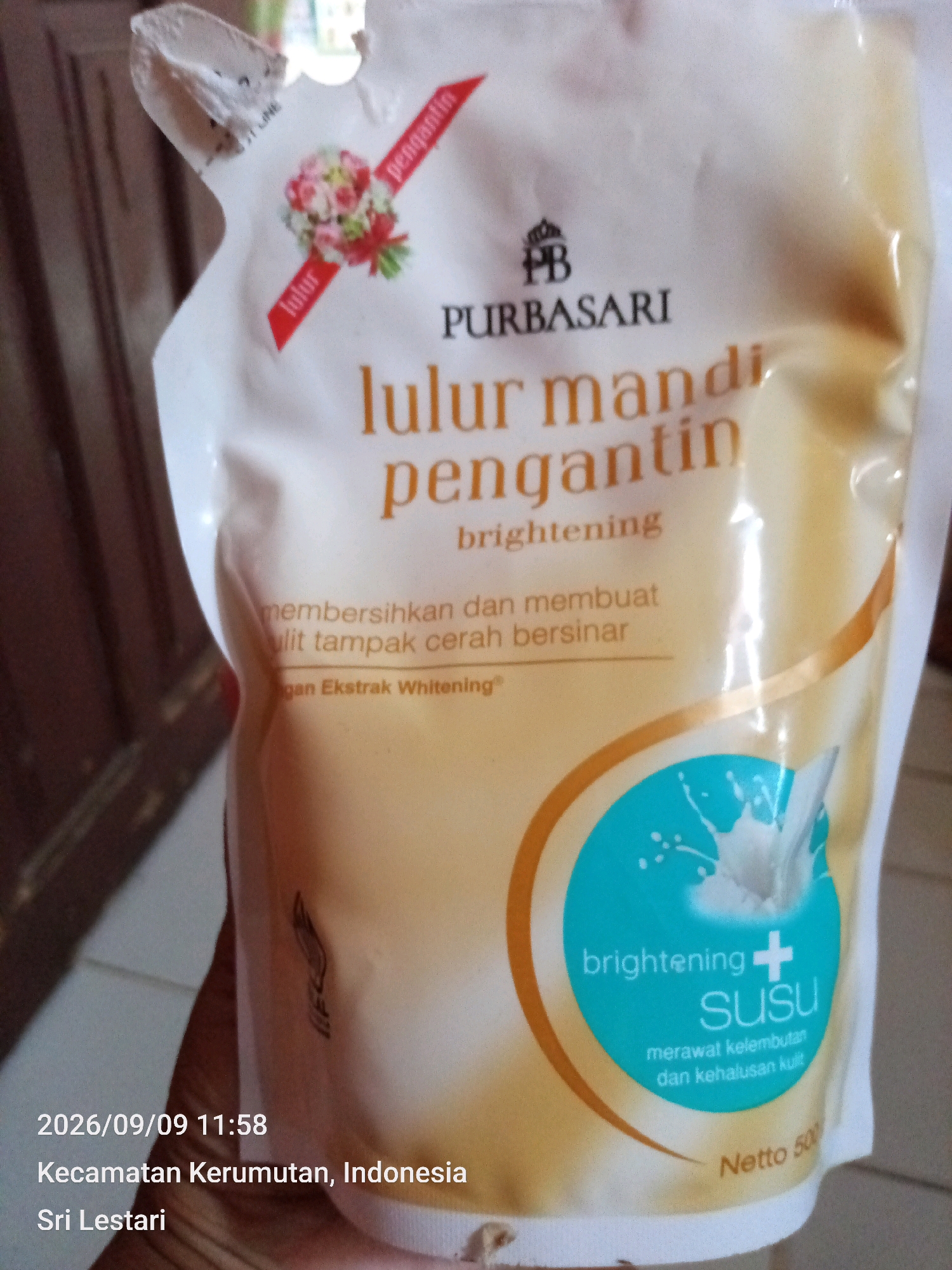 A white pouch of Purbasari Lulur Mandi Pengantin brightening body scrub is held by a person's hand. The pouch has an illustration of a bouquet of flowers and text in Indonesian. The text reads "Lulur Mandi Pengantin brightening" and "membersihkan dan membuat kulit tampak cerah bersinar dengan Ekstrak Whitening". A circular graphic on the pouch contains the words "brightening SUSU" and "merawat kelembutan dan kehalusan kulit". The pouch also displays "Netto 500g". The date and location "2026/09/09 11:58, Kecamatan Kerumutan, Indonesia, Sri Lestari" are superimposed on the bottom left of the image. The background appears to be a room with wooden furniture.