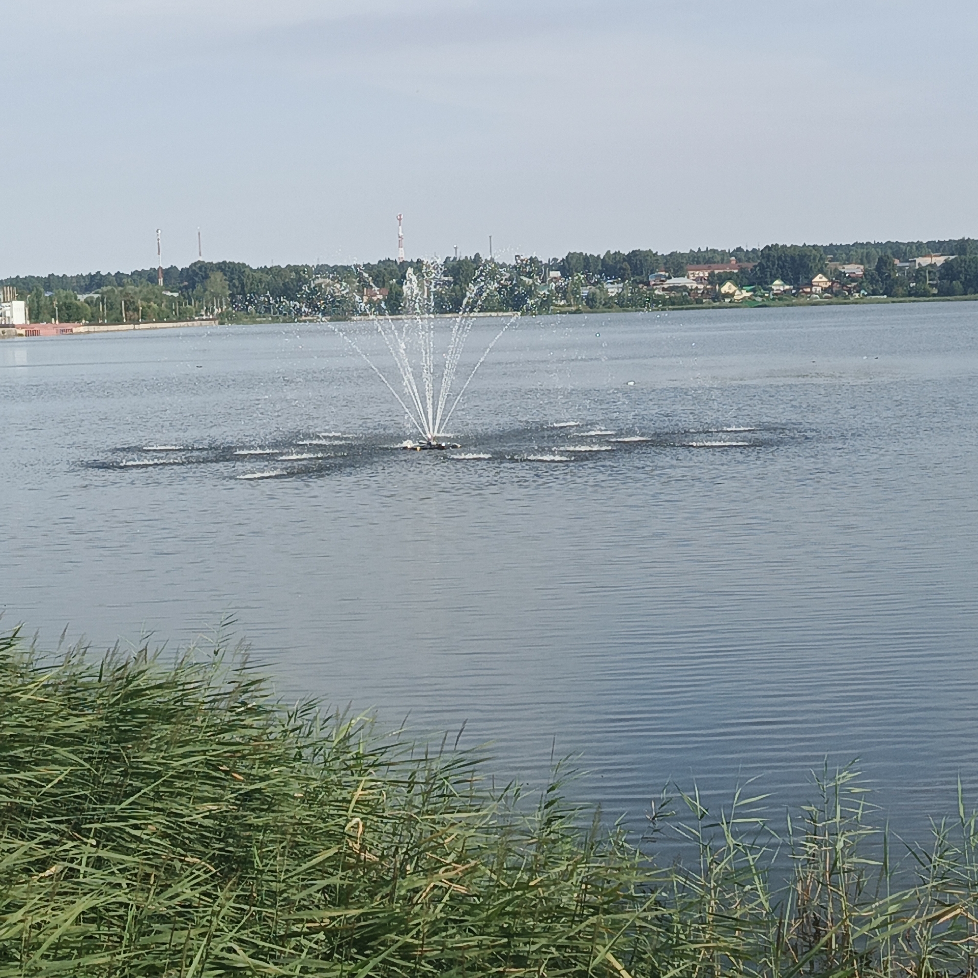 A body of water, likely a lake, is the primary focus of the image. A fountain sprays water high into the air in the center of the lake, creating a striking visual. The background depicts a shoreline with a dense line of trees and a few scattered buildings, suggesting a populated area on the opposite bank. In the foreground, tall green reeds grow along the edge of the water. The sky above is a pale blue with light clouds. The scene appears to be in the city of Kambarka, Russia, given the presence of industrial structures and the distinctive landscape.