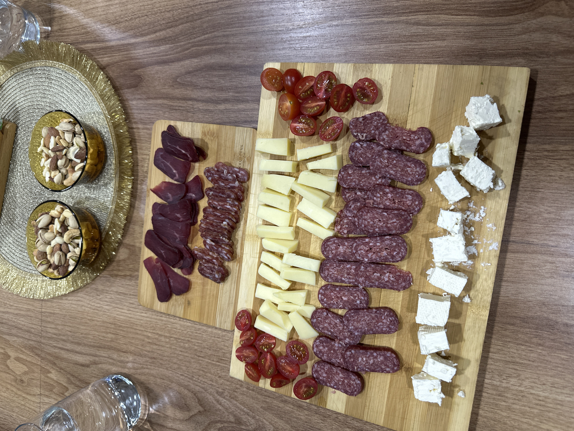 The image shows two wooden cutting boards arranged on a wooden table. On the left, a smaller board displays slices of dried beef and thin slices of sausage. To the right, a larger board contains cherry tomatoes, slices of yellow cheese, salami, and cubes of white cheese. Two small bowls filled with mixed nuts sit on a golden placemat to the left of the boards. The location, Lehchevo, Bulgaria is not present in the image.